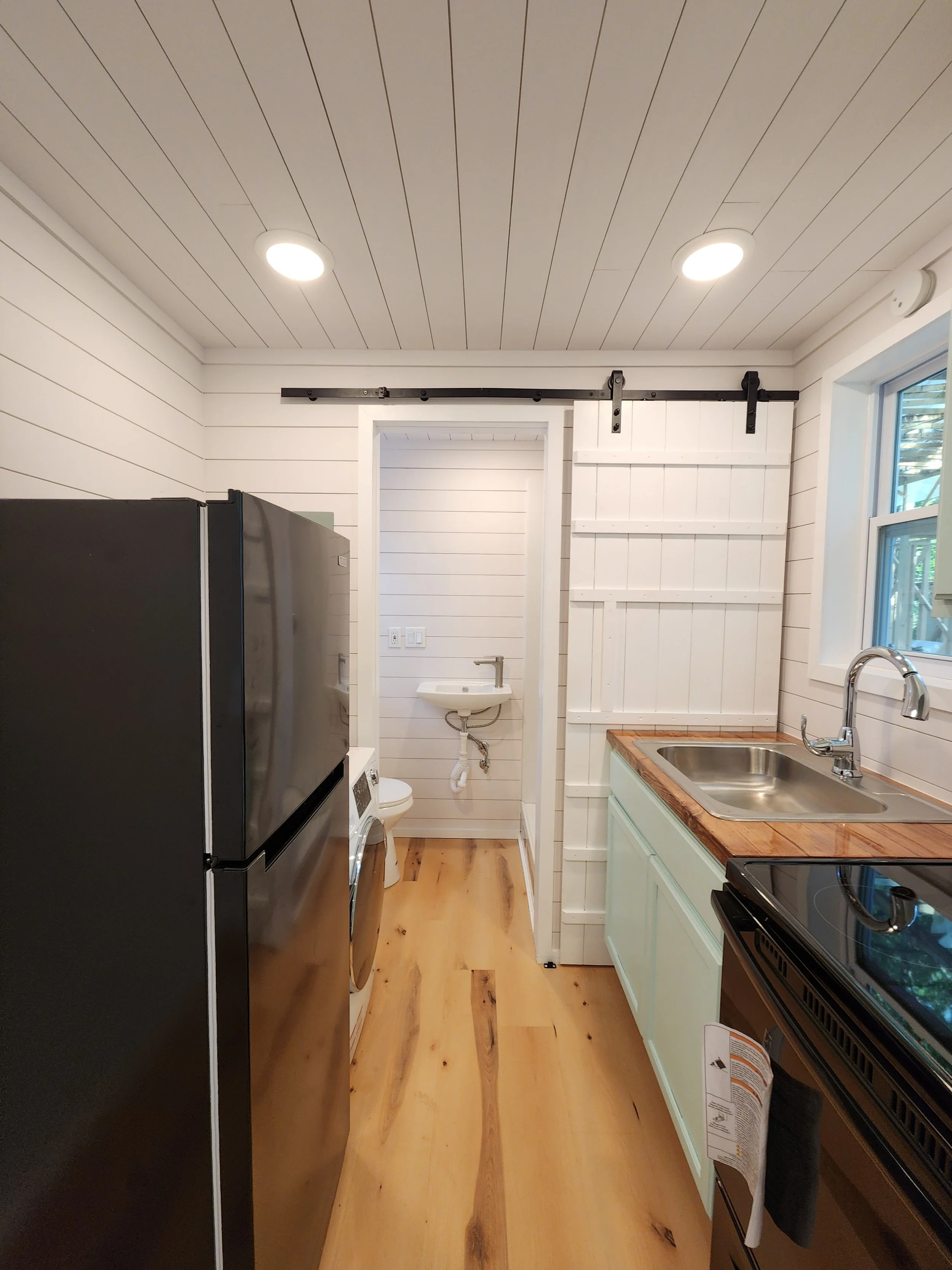 A small kitchen with light green cabinets, a stainless steel sink, black stove, and a refrigerator. In the background, there is a small bathroom with a toilet and a wall-mounted sink, separated by a sliding barn door. The room has white shiplap walls