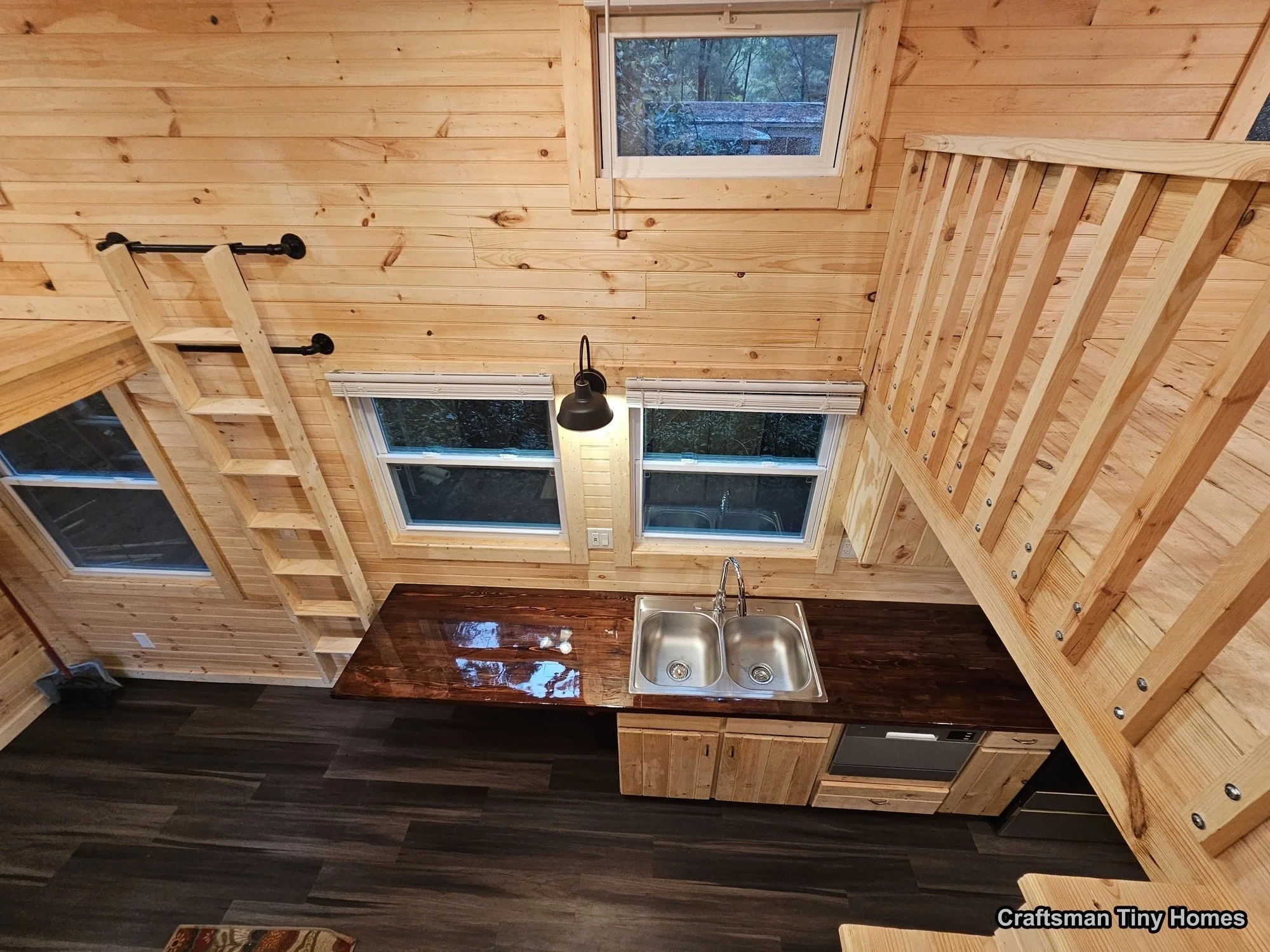 Interior of a tiny home kitchen with wooden walls, a dark wood countertop, a double stainless steel sink, three windows, a black wall-mounted lamp, a ladder on the left leading up to a loft area, and another ladder on the right with a railing. There 