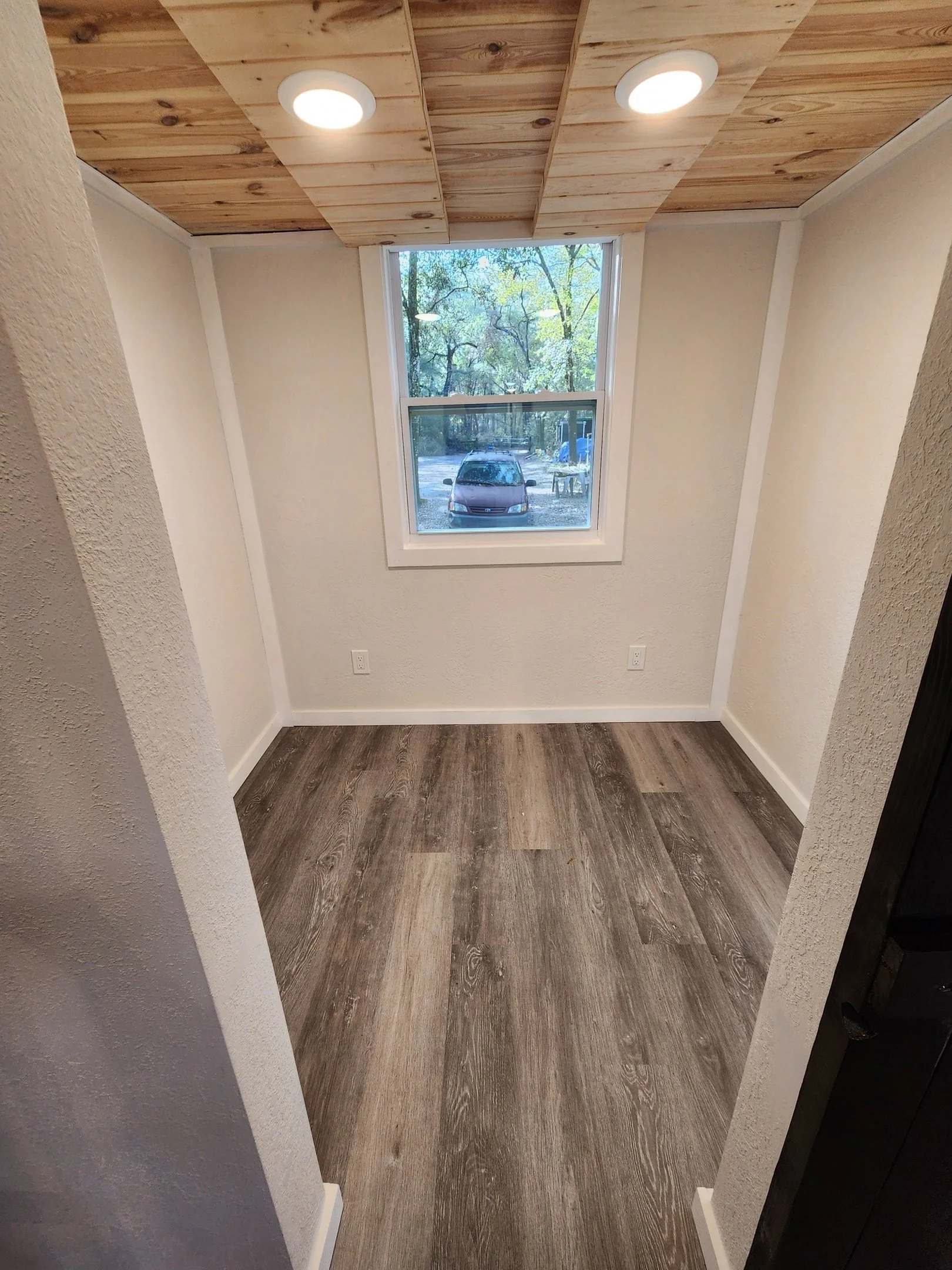 Empty room with wood flooring, white textured walls, a large window showing trees and a parked car outside, a wood-paneled ceiling with recessed lighting.