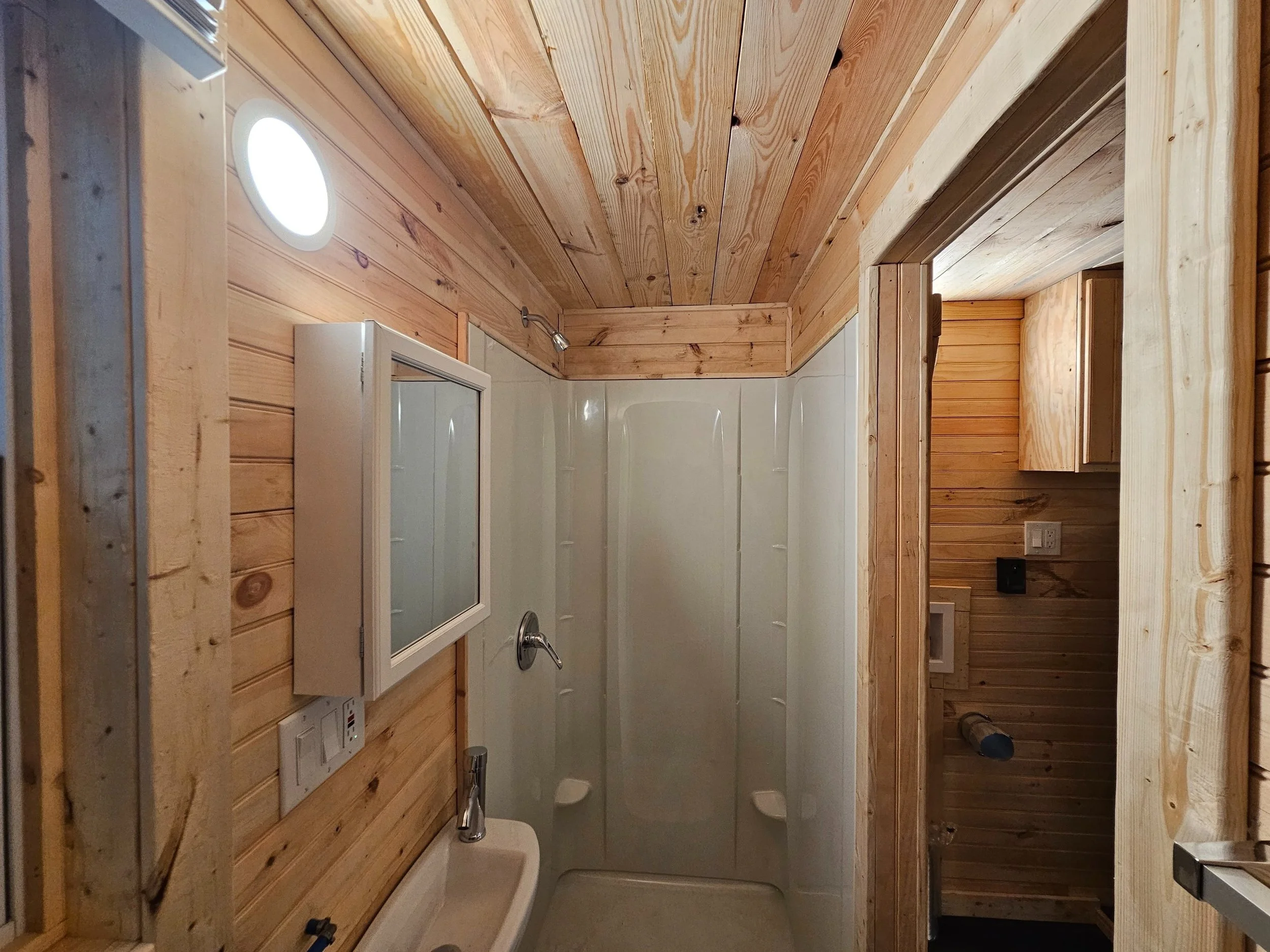Small bathroom with wood-paneled walls and ceiling, shower with frosted glass door, wall-mounted mirror cabinet, small sink with faucet, and partially visible toilet in an adjacent room.