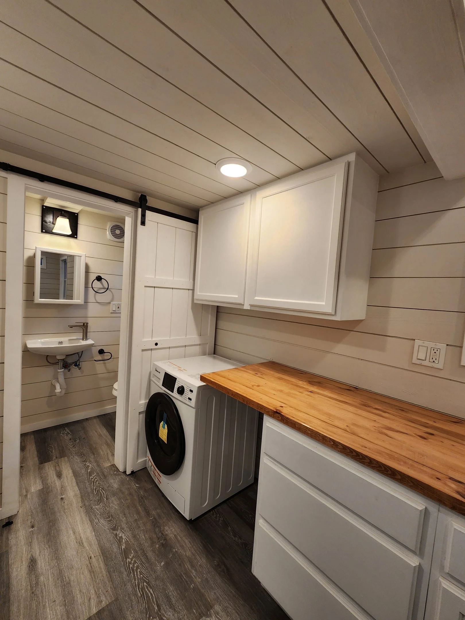 Laundry room with a washing machine, white cabinetry, a wooden countertop, and a small bathroom with a wall-mounted sink and mirror.