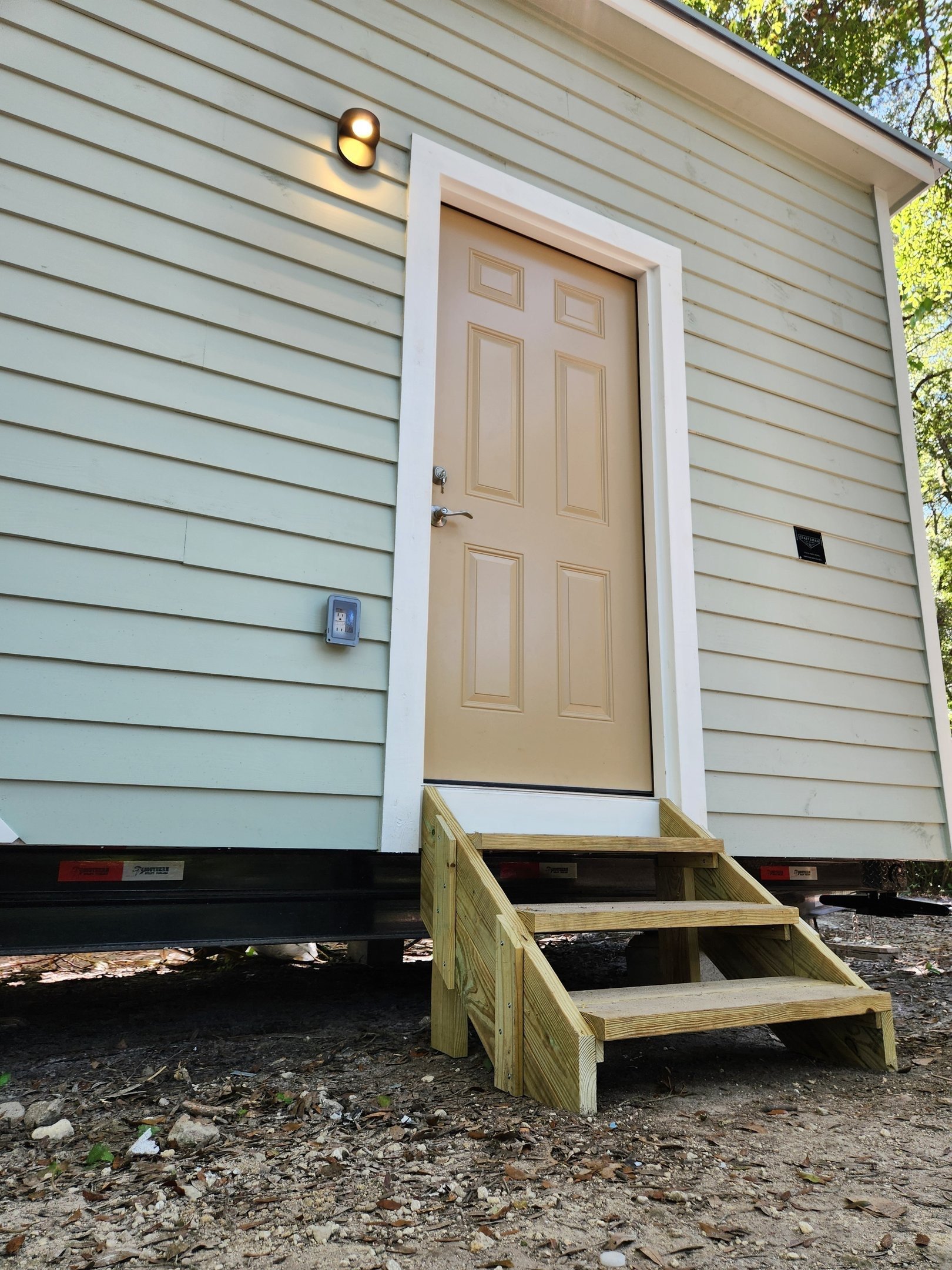 Small beige shed with a staircase leading to a door, situated on uneven ground, with siding and a light fixture outside.