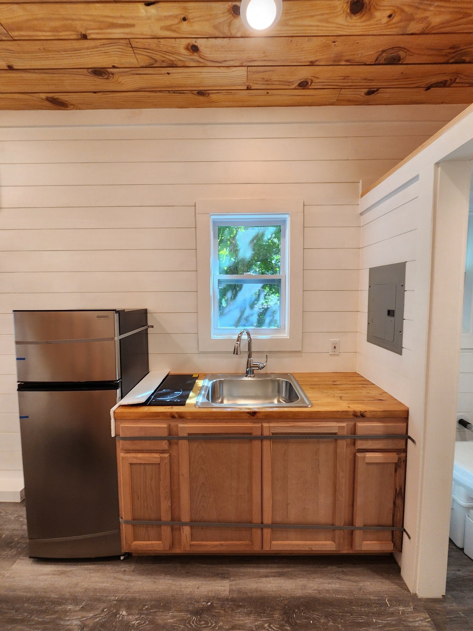 Small kitchen with wooden cabinets, stainless steel refrigerator, small sink, window above the sink, and a wooden ceiling.