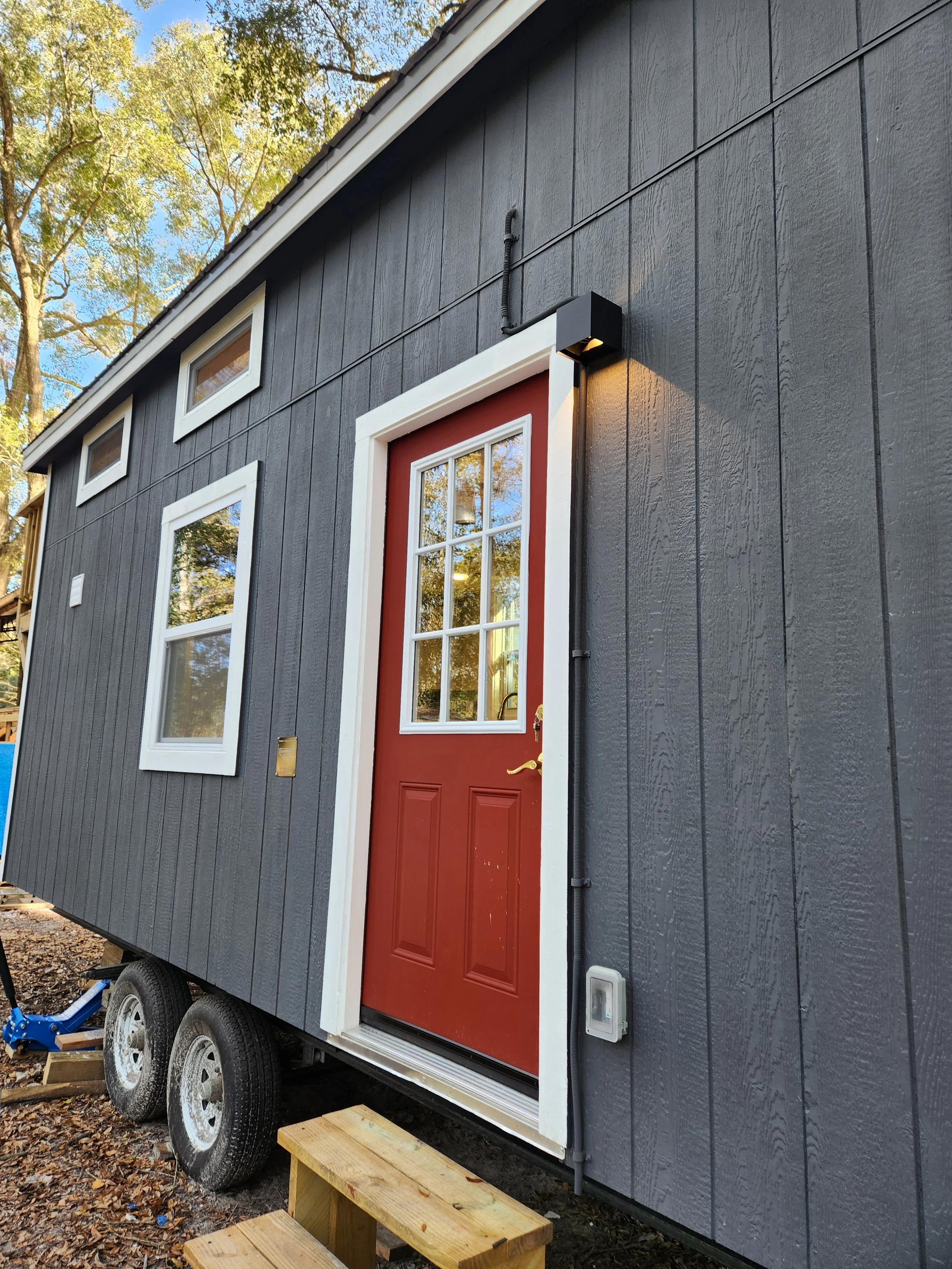A tiny house on wheels with dark gray exterior siding, a red front door with glass panels, three small horizontal windows at the top, two larger windows on the side, outdoor lighting, and a small wooden step.
