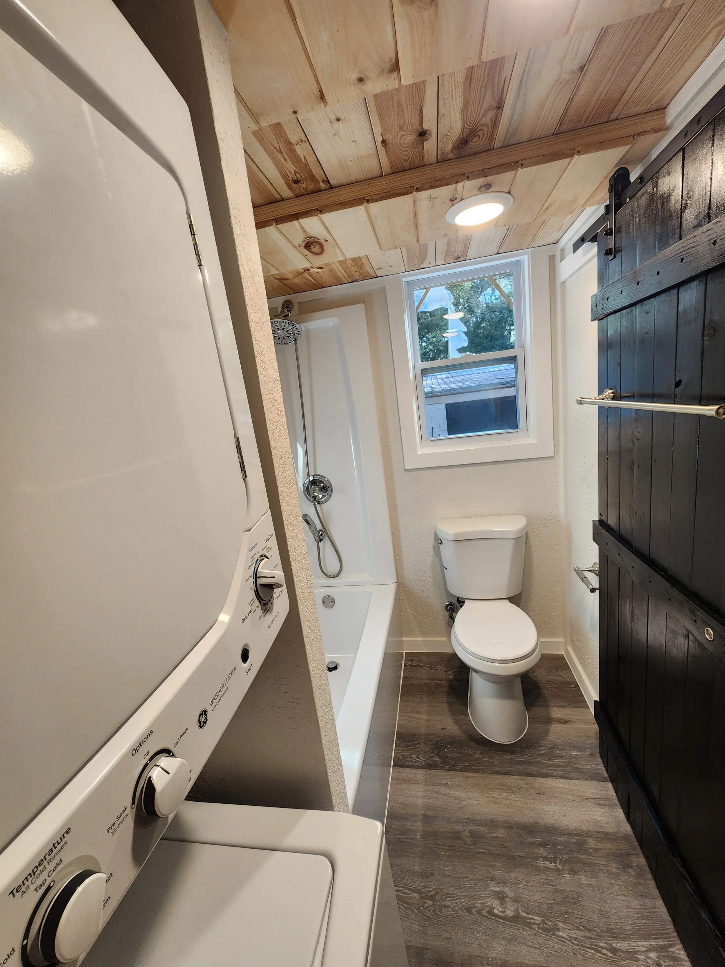 Small bathroom with wooden ceiling, white walls, a window showing trees outside, a white bathtub with a showerhead, a white toilet, a black cabinet, and a washing machine with dials visible.
