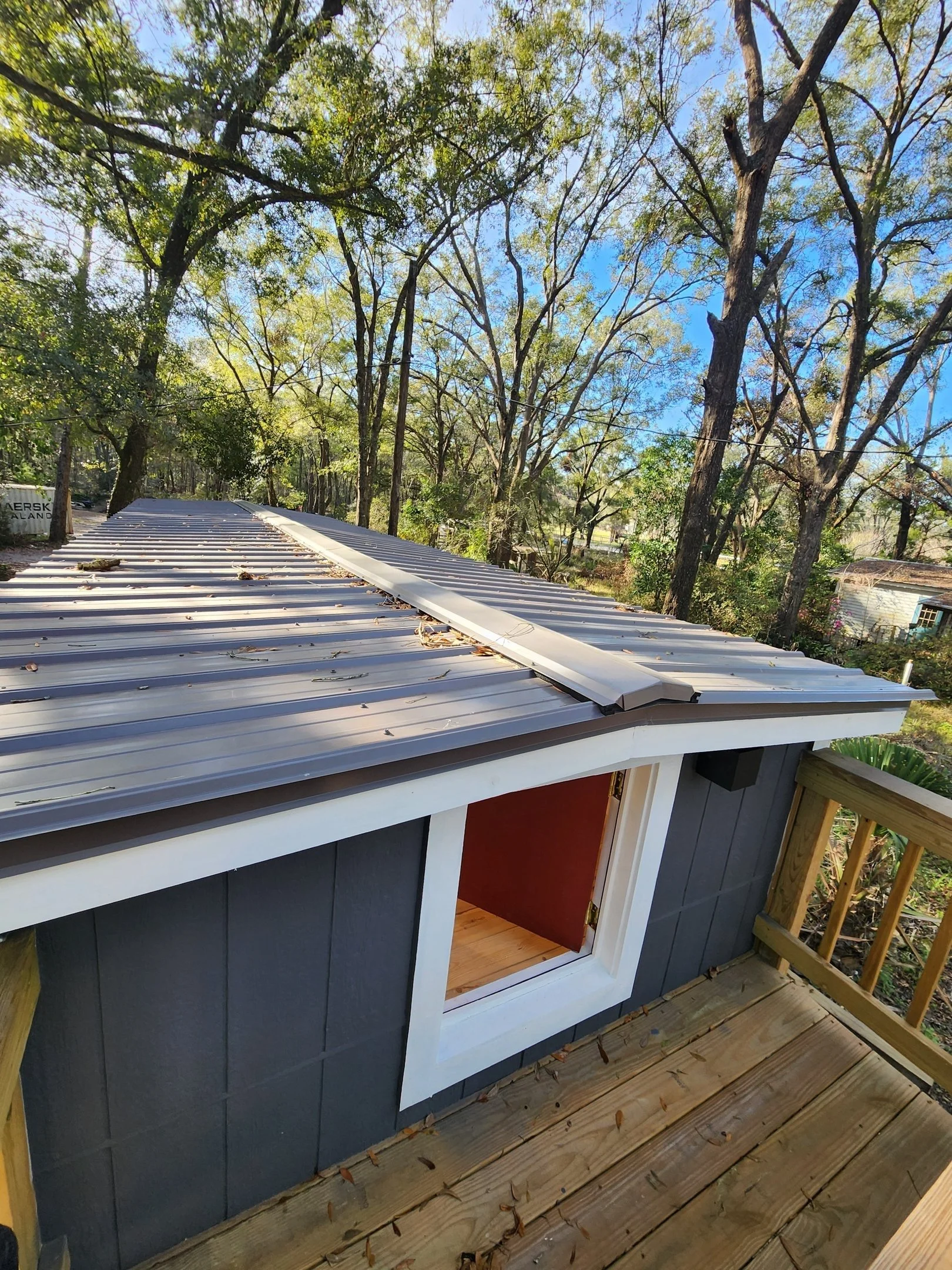 View of a small shed with a metal roof and an open window, with a wooden deck and railing. The background shows trees with green leaves and a blue sky.