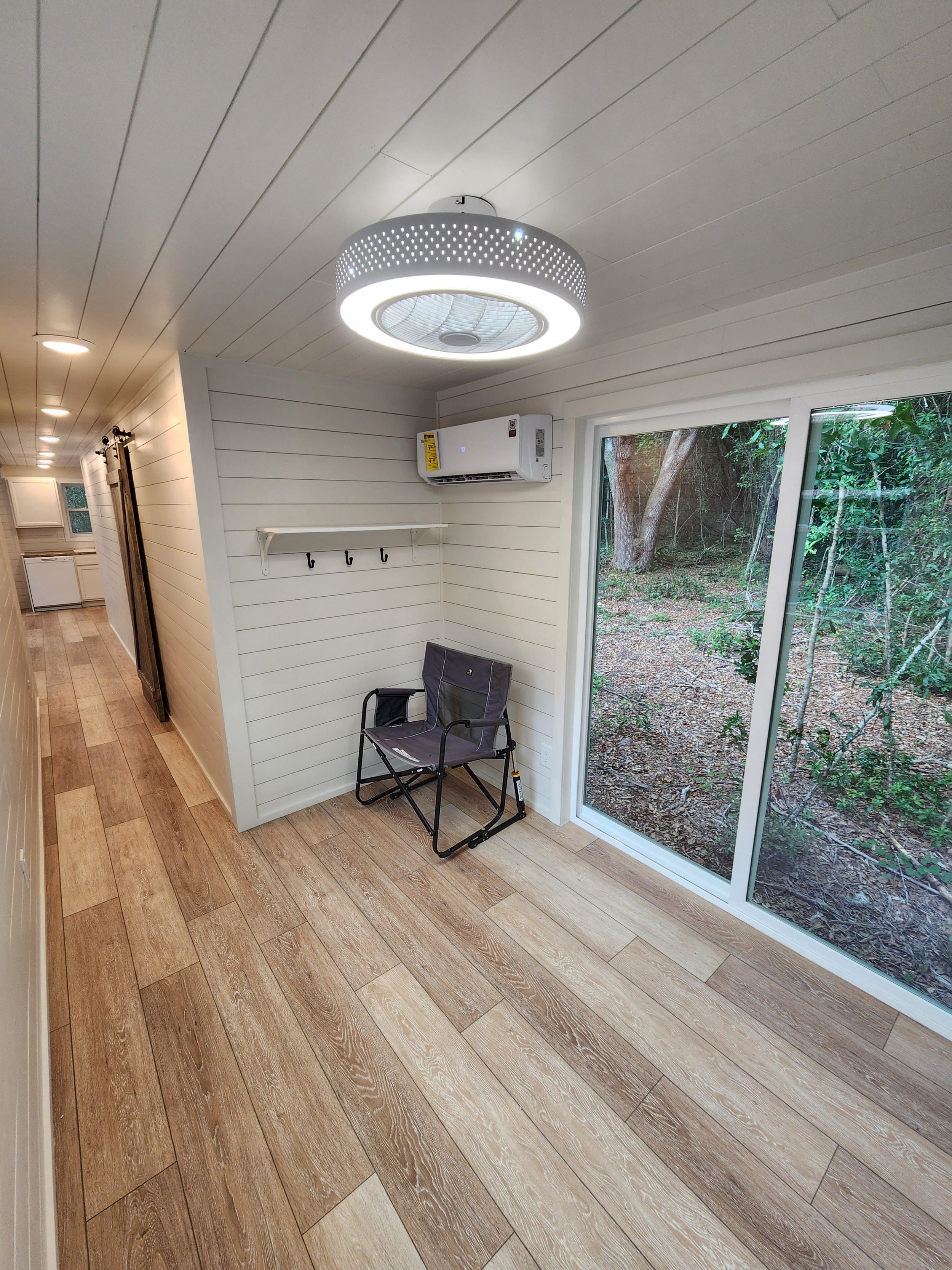 A small room with a wooden floor, white shiplap walls, and a ceiling-mounted circular light fixture. There is a wall-mounted air conditioner, a white shelf with hooks, a folding chair, and a large sliding glass door that opens to a wooded outdoor are