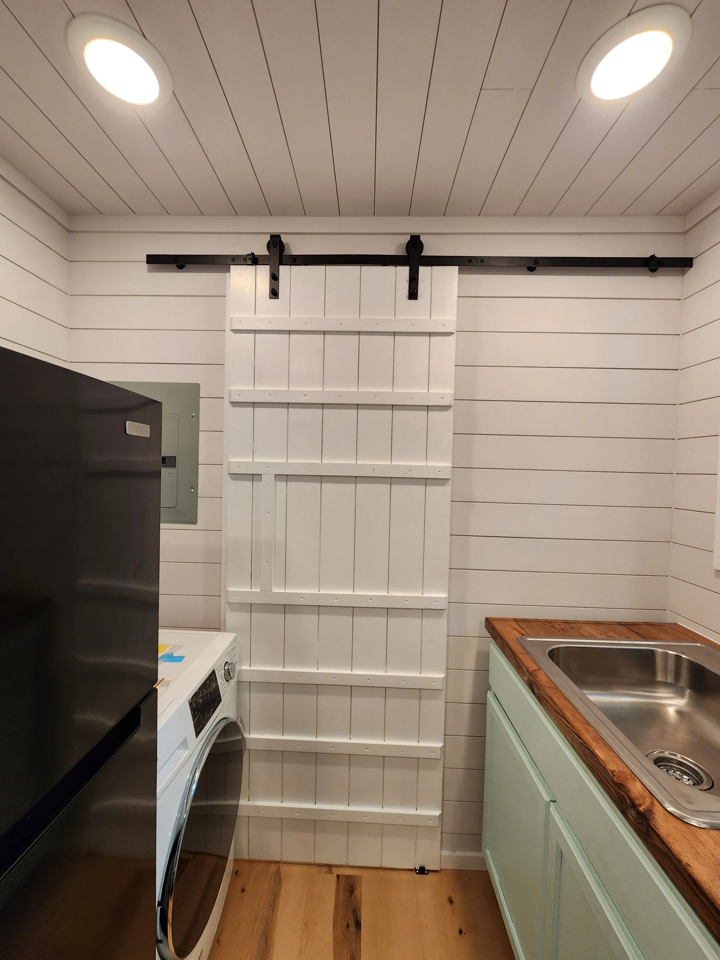 Interior of a laundry room converted into a pantry, with a sliding barn door on a ceiling-mounted track, washer, sink with wooden countertop, and shiplap walls.