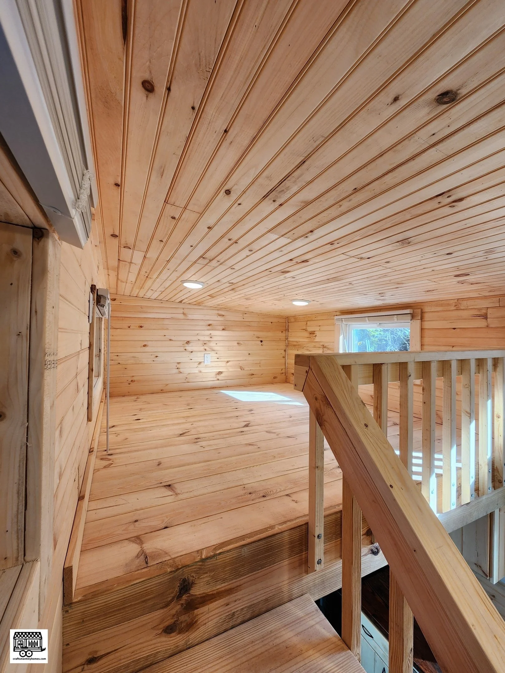 Interior of a small wooden room or loft with wooden planks on the ceiling and walls, a window letting in natural light, and a wooden railing around a staircase opening.