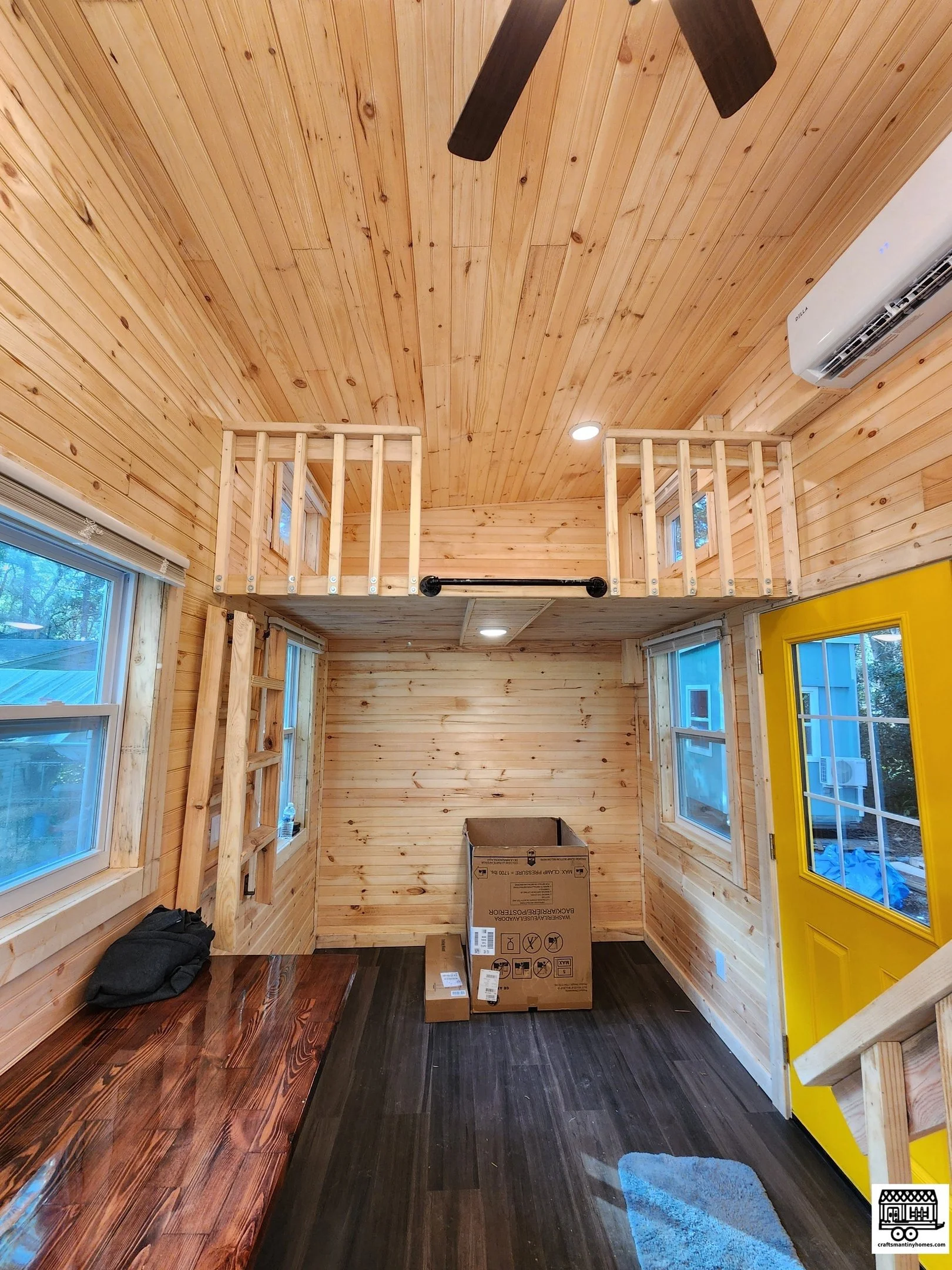 Interior of a small wooden room with a loft. The room has wood paneling for the walls and ceiling, three windows, a bright yellow door, a black ceiling fan, and an air conditioning unit. There's a wooden bench against the wall, a cardboard box on the
