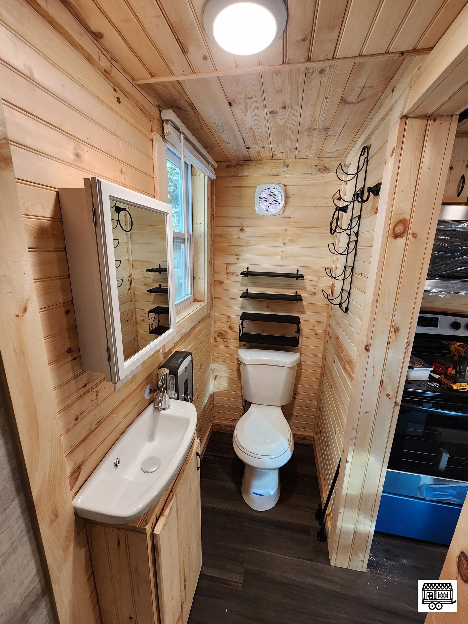 Small rustic bathroom with wood-paneled walls and ceiling, featuring a white toilet, a small wall-mounted sink, a mirror cabinet, a window, and wall-mounted shelves and accessories.