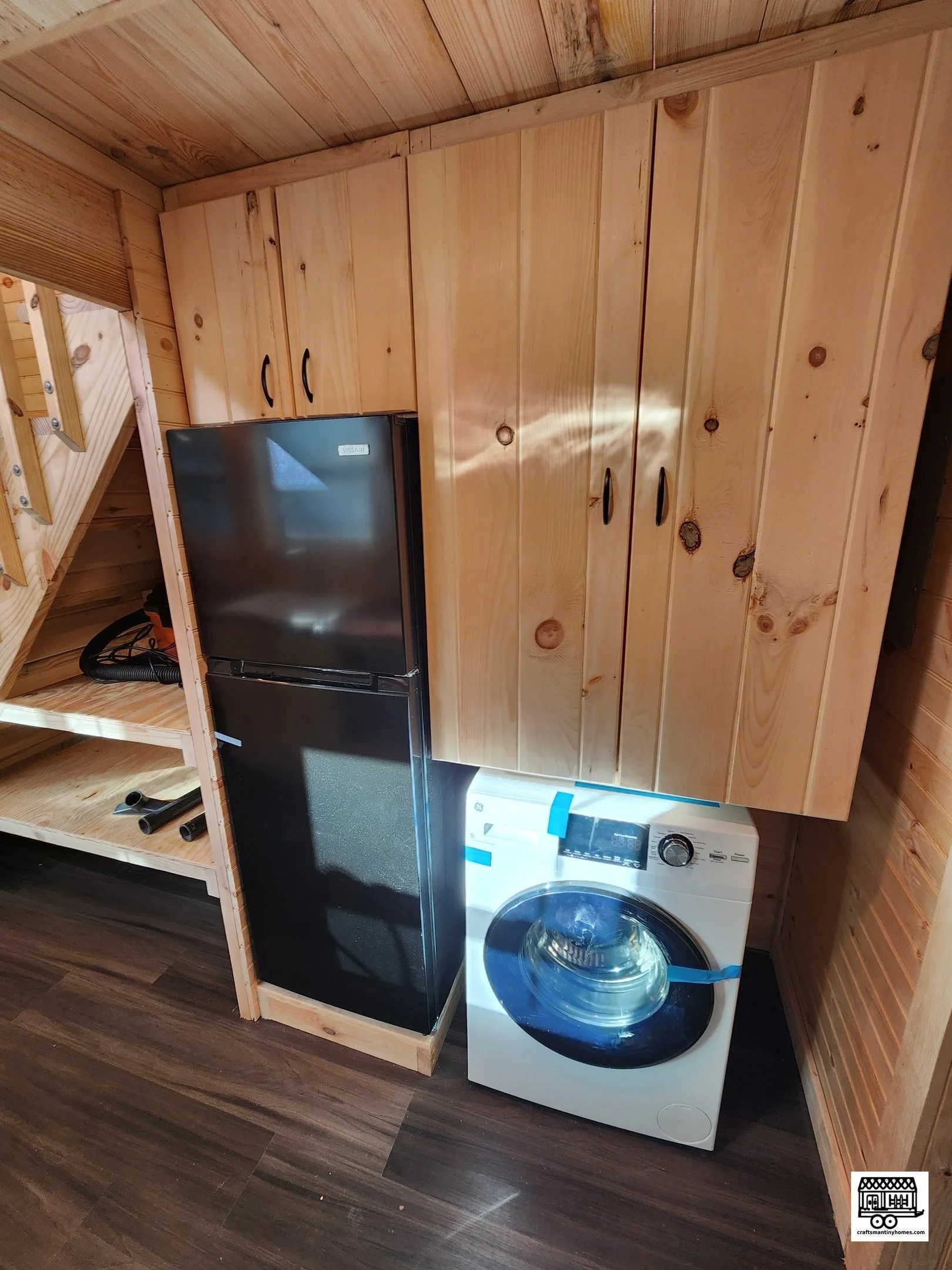 A small black refrigerator and a white front-loading washing machine in a wooden room with light-colored wood paneling on walls and ceiling, and dark wood flooring.