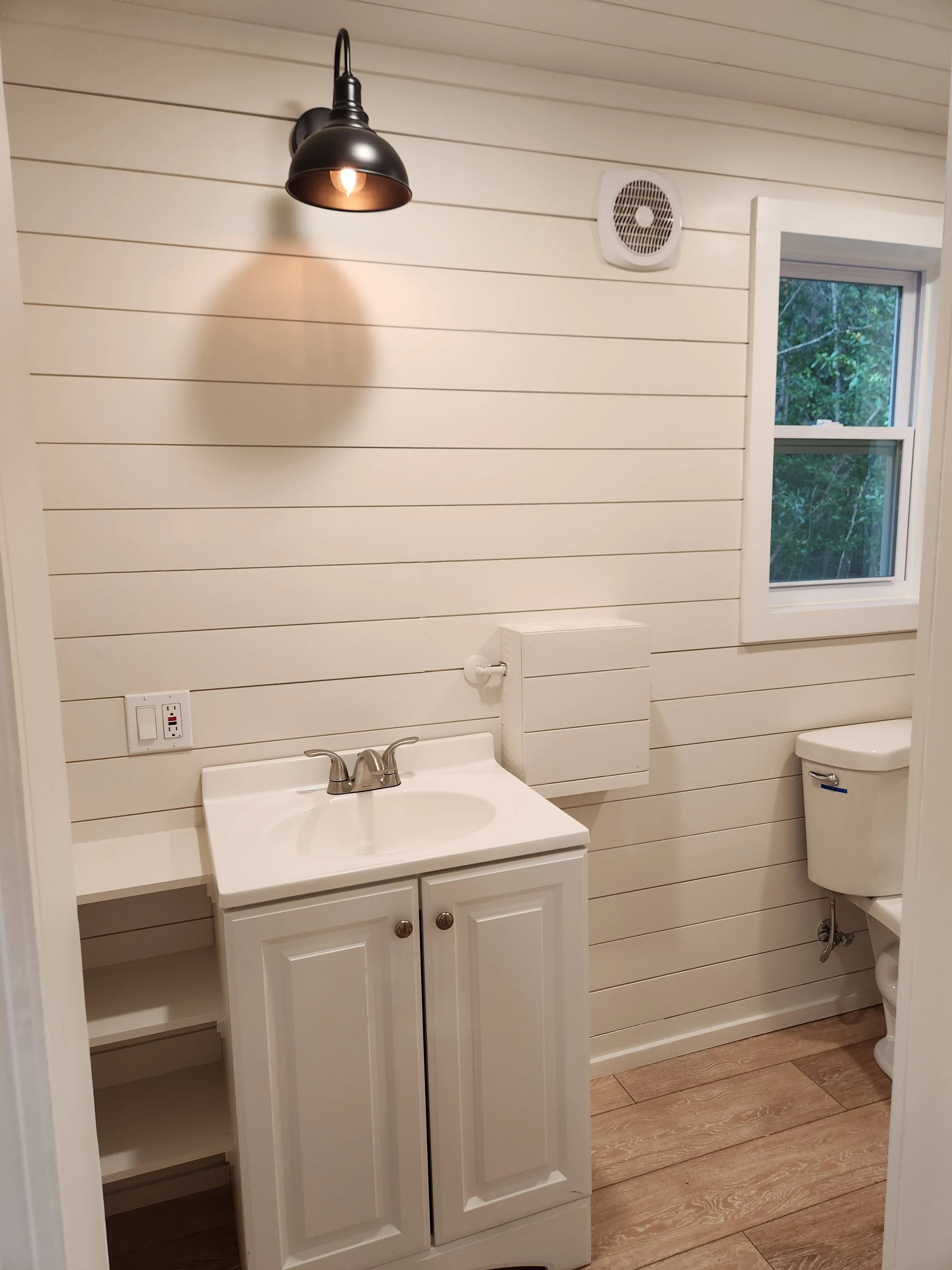 A small bathroom with white shiplap walls, a black wall-mounted light fixture, a white vanity with a sink, a wall-mounted toilet paper holder, a window with trees outside, and a toilet. The bathroom has wooden flooring.