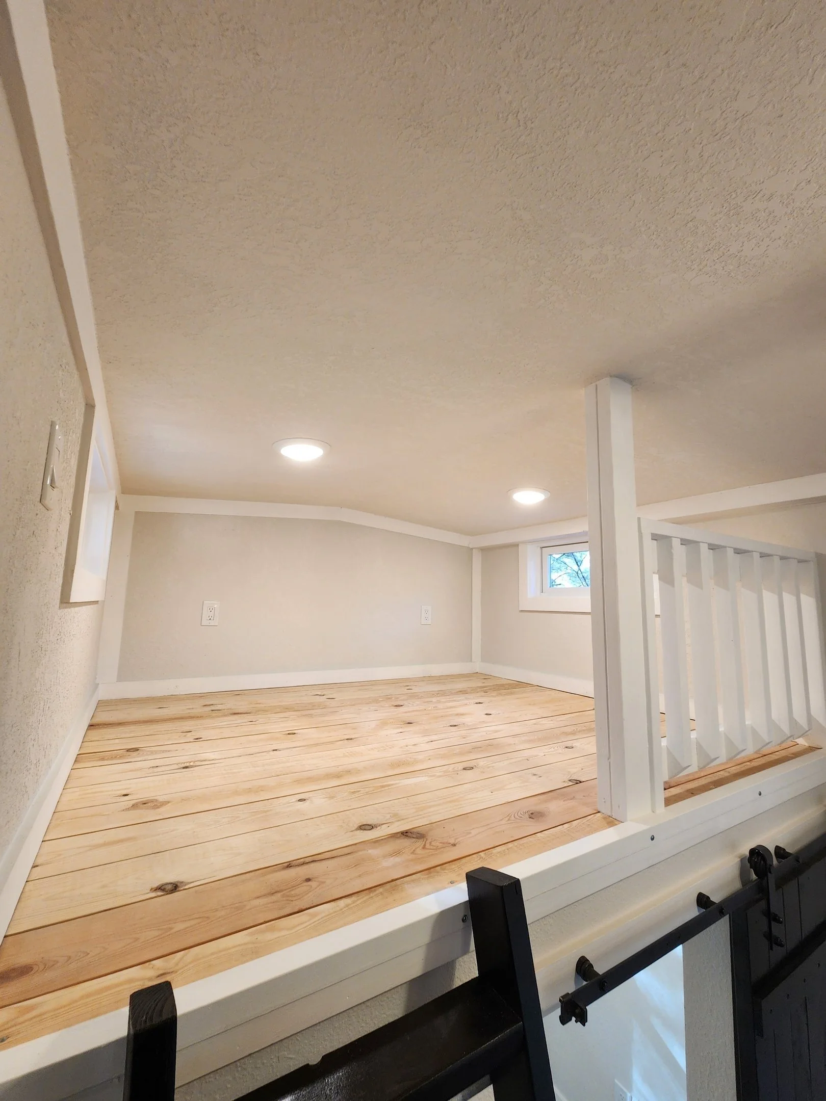 Empty attic space with wooden floor, white walls, small windows, and ceiling lights.