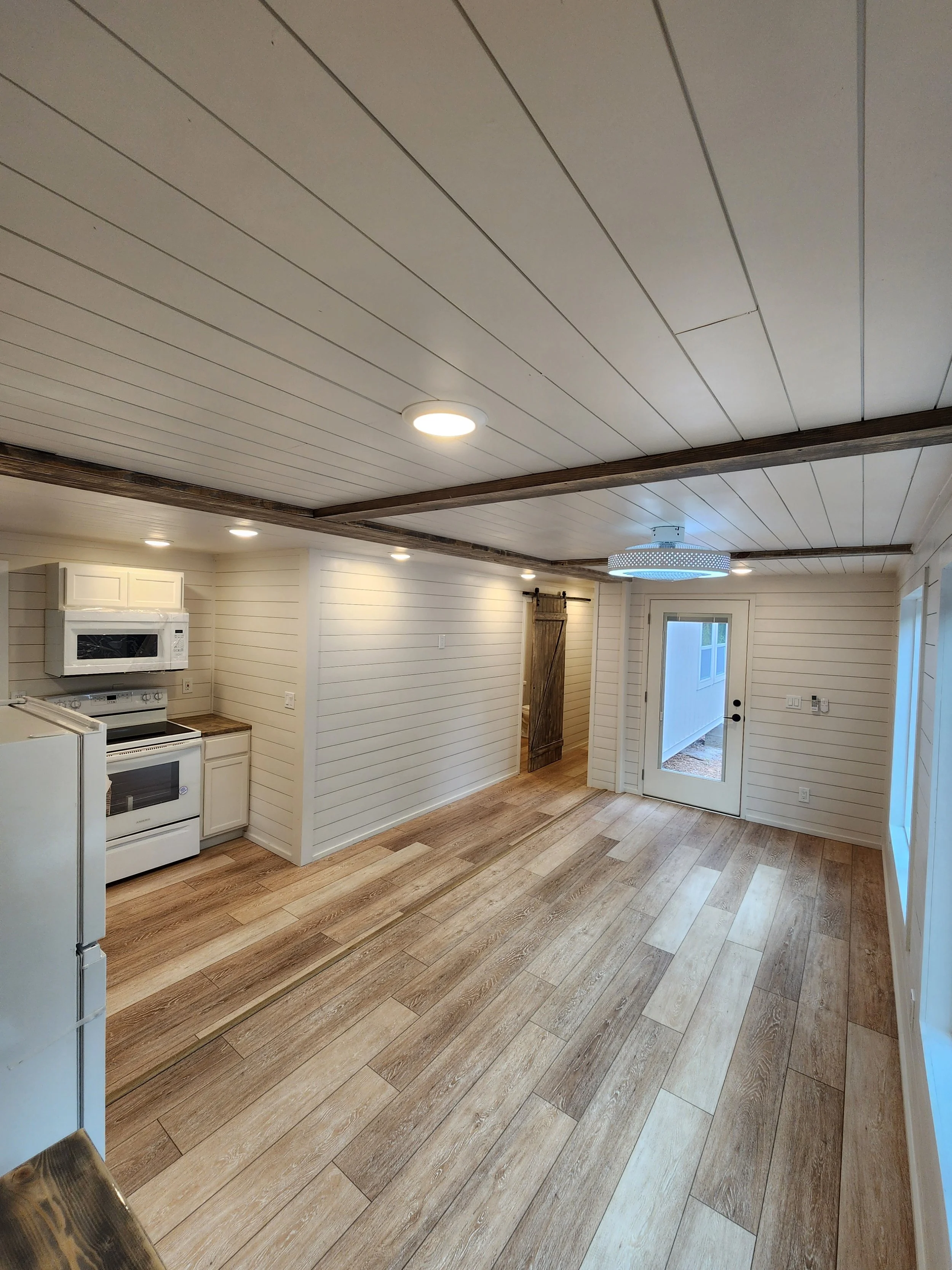 Empty room with wood flooring, white paneled walls, ceiling with recessed lighting, a modern circular ceiling light, kitchen area with stove, microwave, and refrigerator, and a door leading outside.