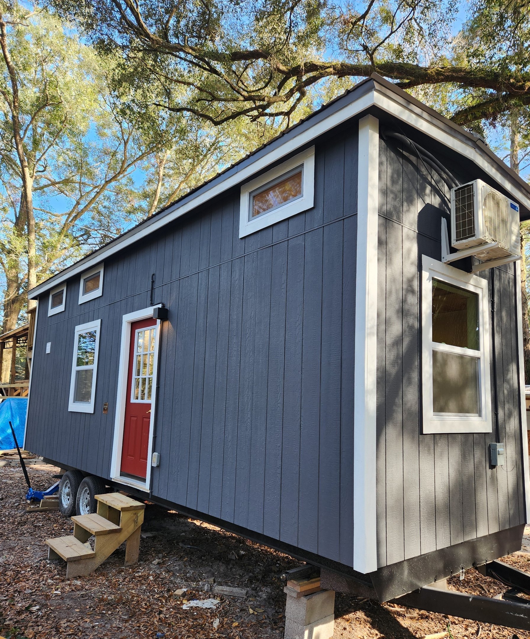 A tiny house on wheels with dark gray vertical siding, white trim, and a red door, set on concrete blocks, surrounded by trees with a clear blue sky above.