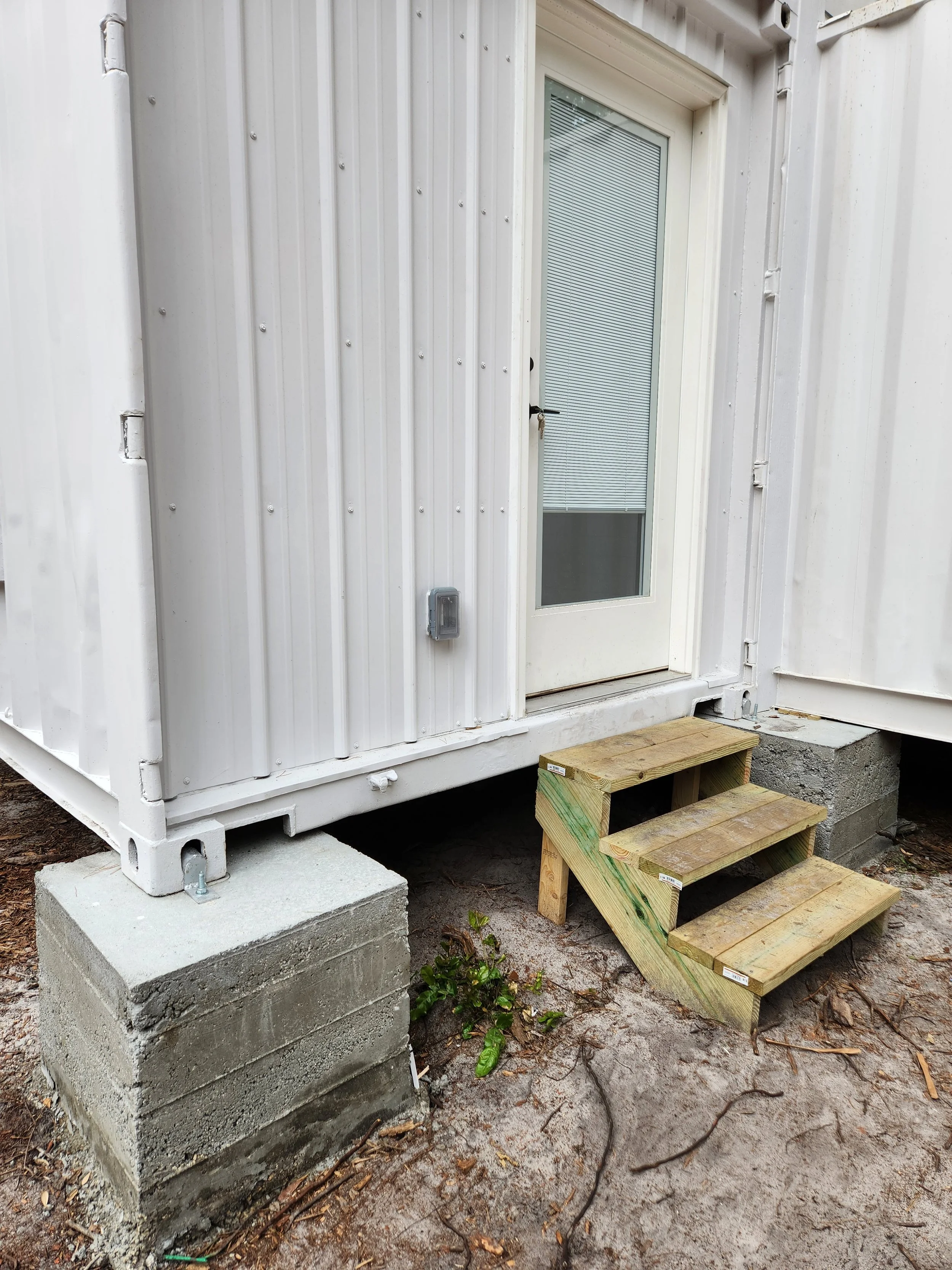 Small wooden stairs leading up to a glass door with a lock. The door is part of a white metal building with a small exterior light and concrete blocks supporting the structure. There is some dirt and a small plant on the ground nearby.