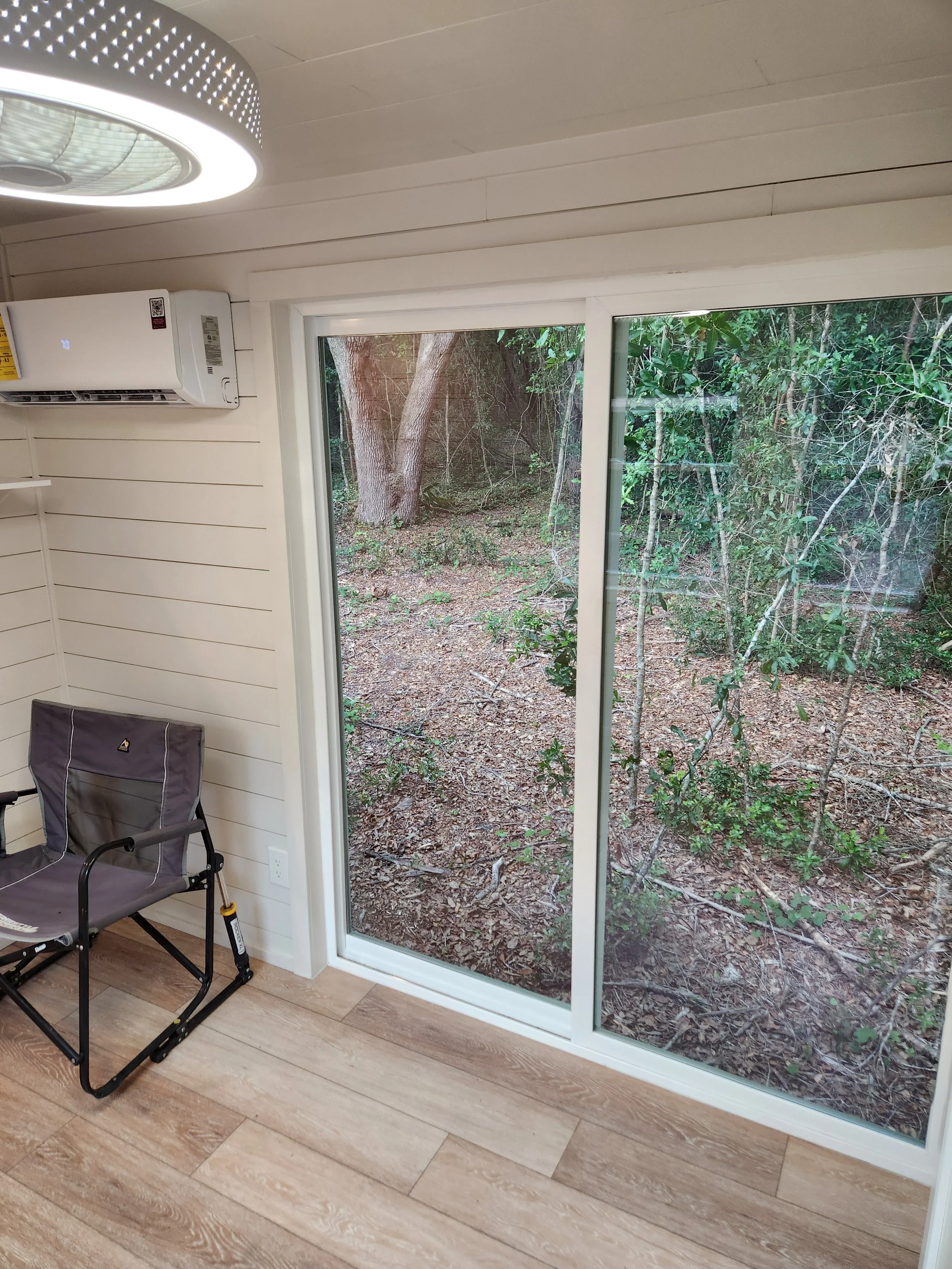 Interior of a room with white paneled walls, a sliding glass door overlooking a wooded outdoor area, a portable black chair, and an air conditioning unit mounted on the wall. A ceiling light fixture is visible.