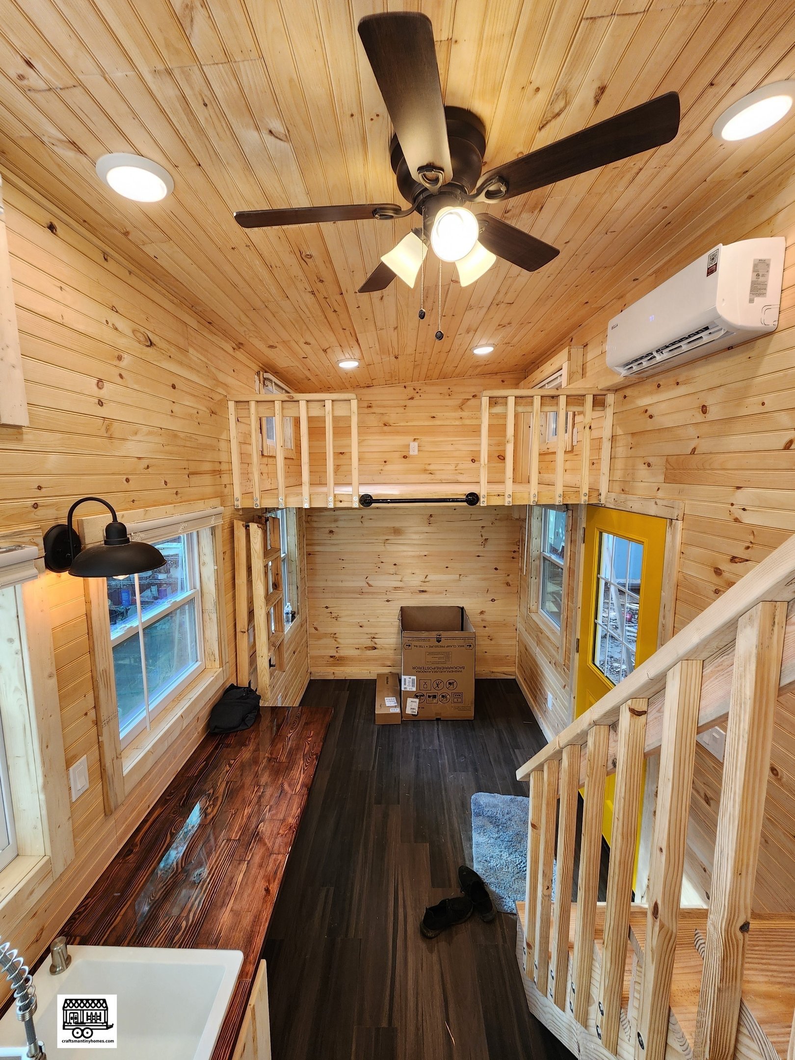 Interior of a tiny house with wood-paneled walls and ceiling, a ceiling fan, windows, a yellow door, a wooden staircase, a wooden table, and a cardboard box.