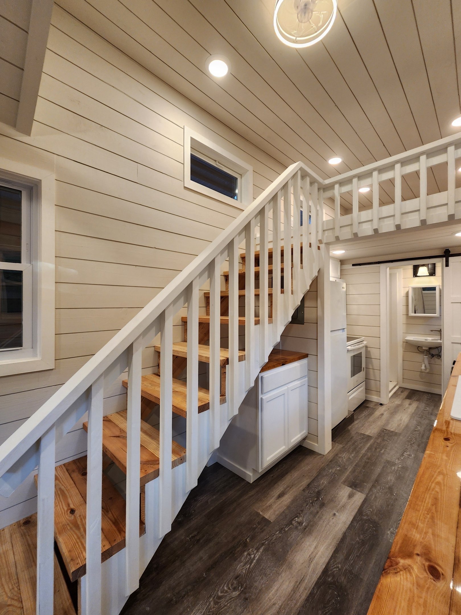 Interior of a house with a wooden staircase leading to a loft, wood-paneled ceiling, white walls with windows, and kitchen appliances.