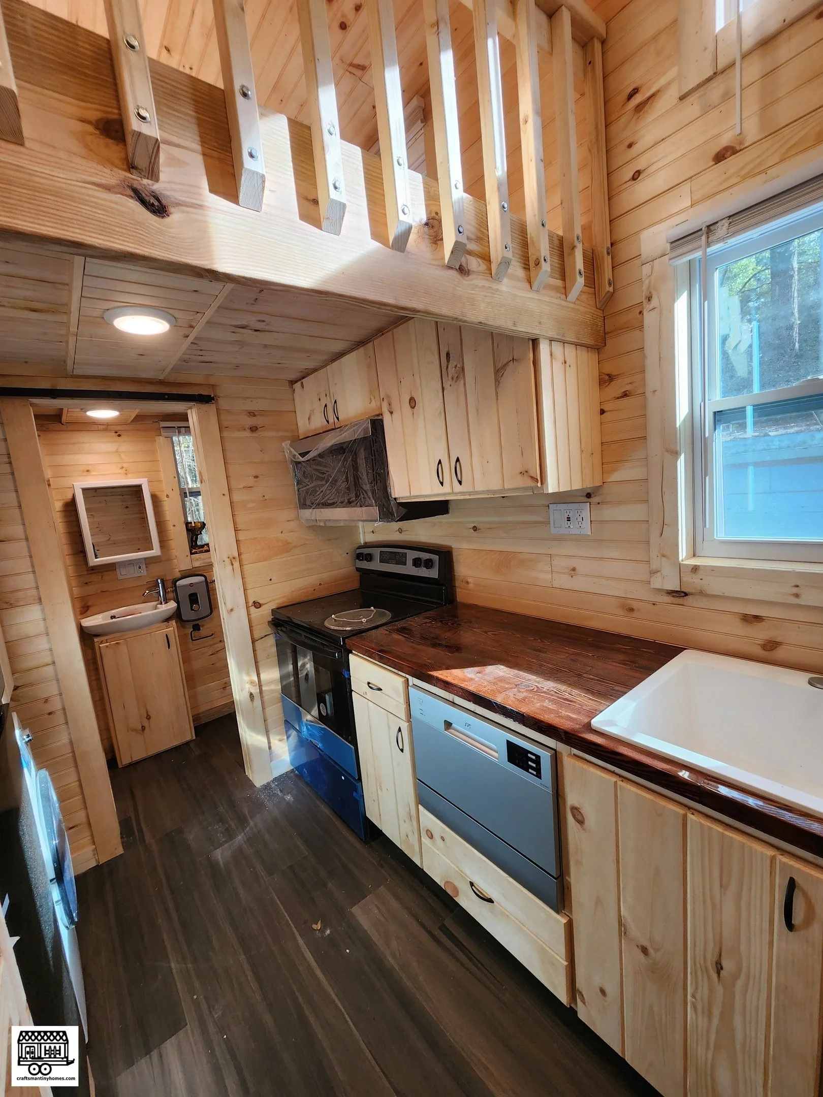 Kitchen with wood-paneled walls, cabinets, and ceiling, a window with a view outside, and a dark wooden countertop. There is a stove, dishwasher, and a sink visible. In the background, a small area with a sink and mirror is seen.