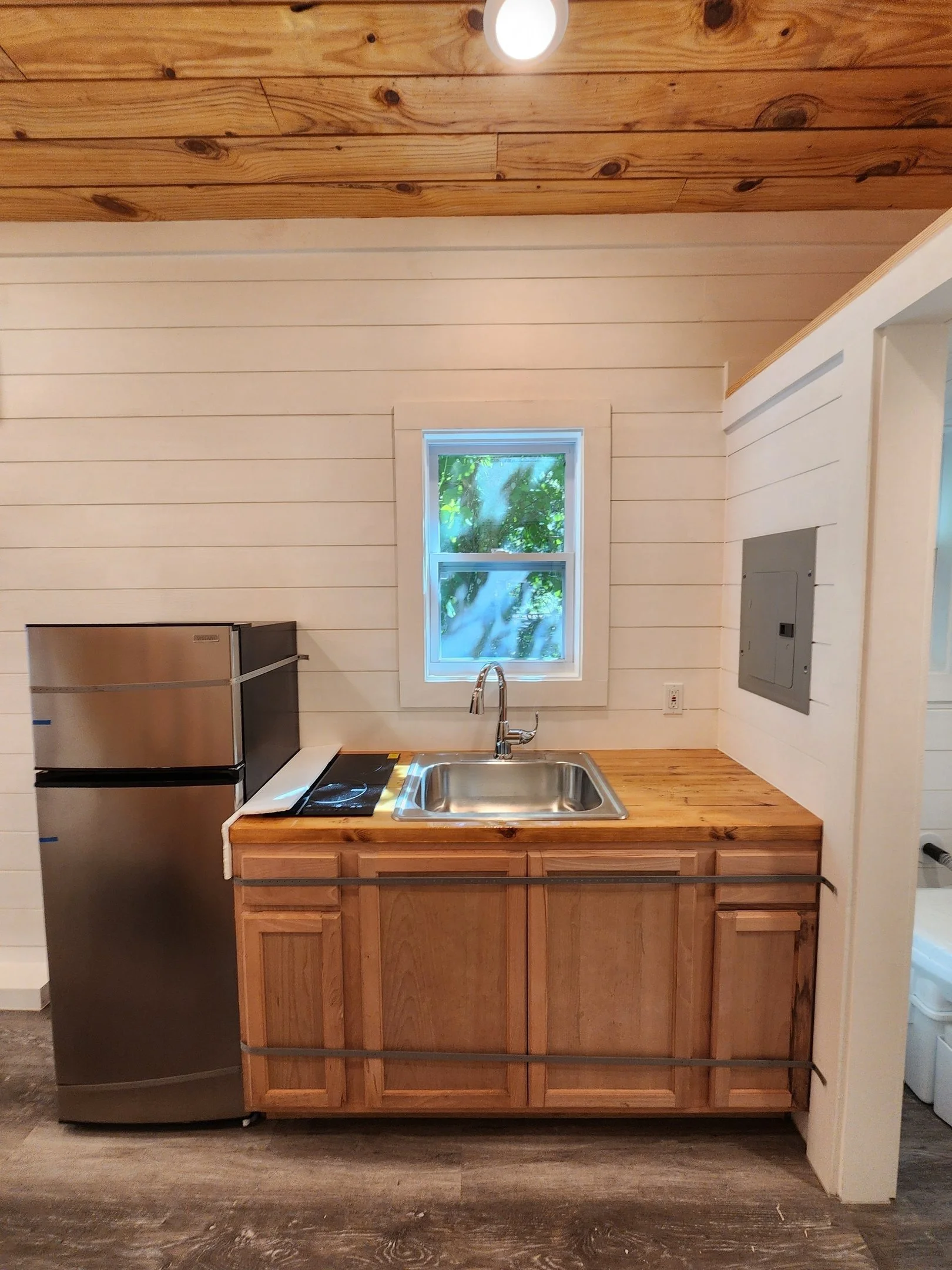 A small kitchen with a wooden countertop, a stainless steel sink, a window with green foliage outside, a stainless steel refrigerator, and a wall panel with an electrical panel.