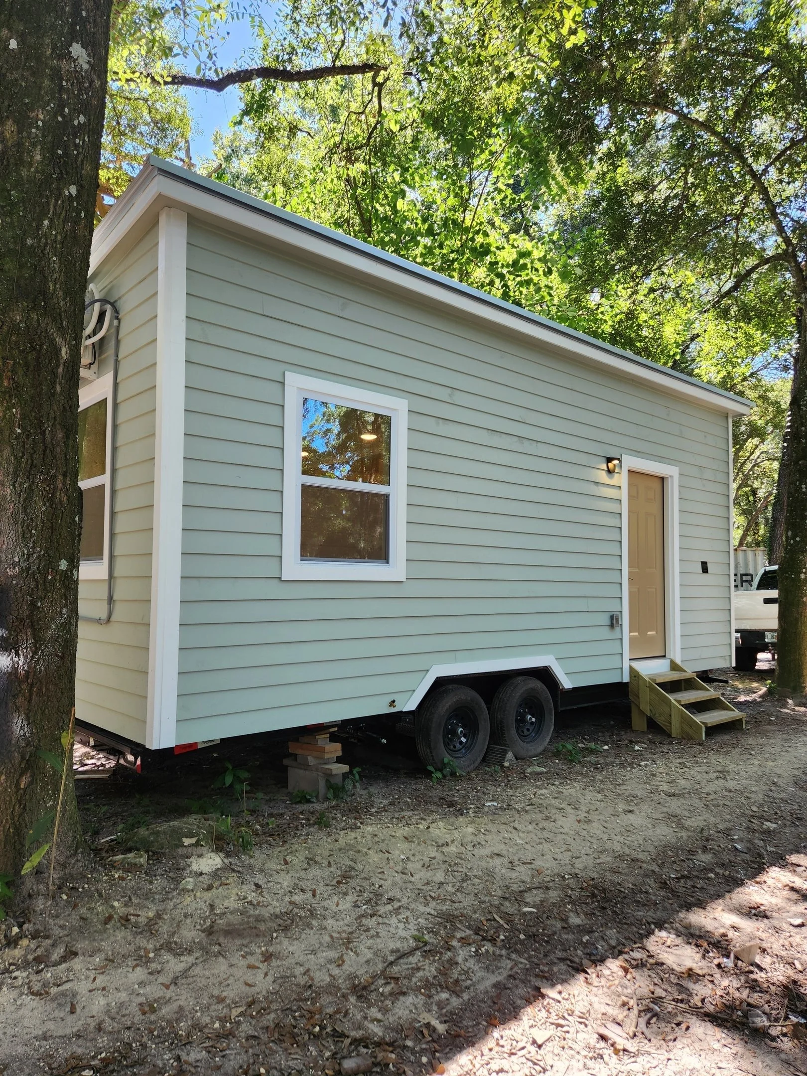 A small, light green mobile home with white trim on a dirt lot, supported on blocks and tires, surrounded by trees, with a window and a door with small wooden steps in front.