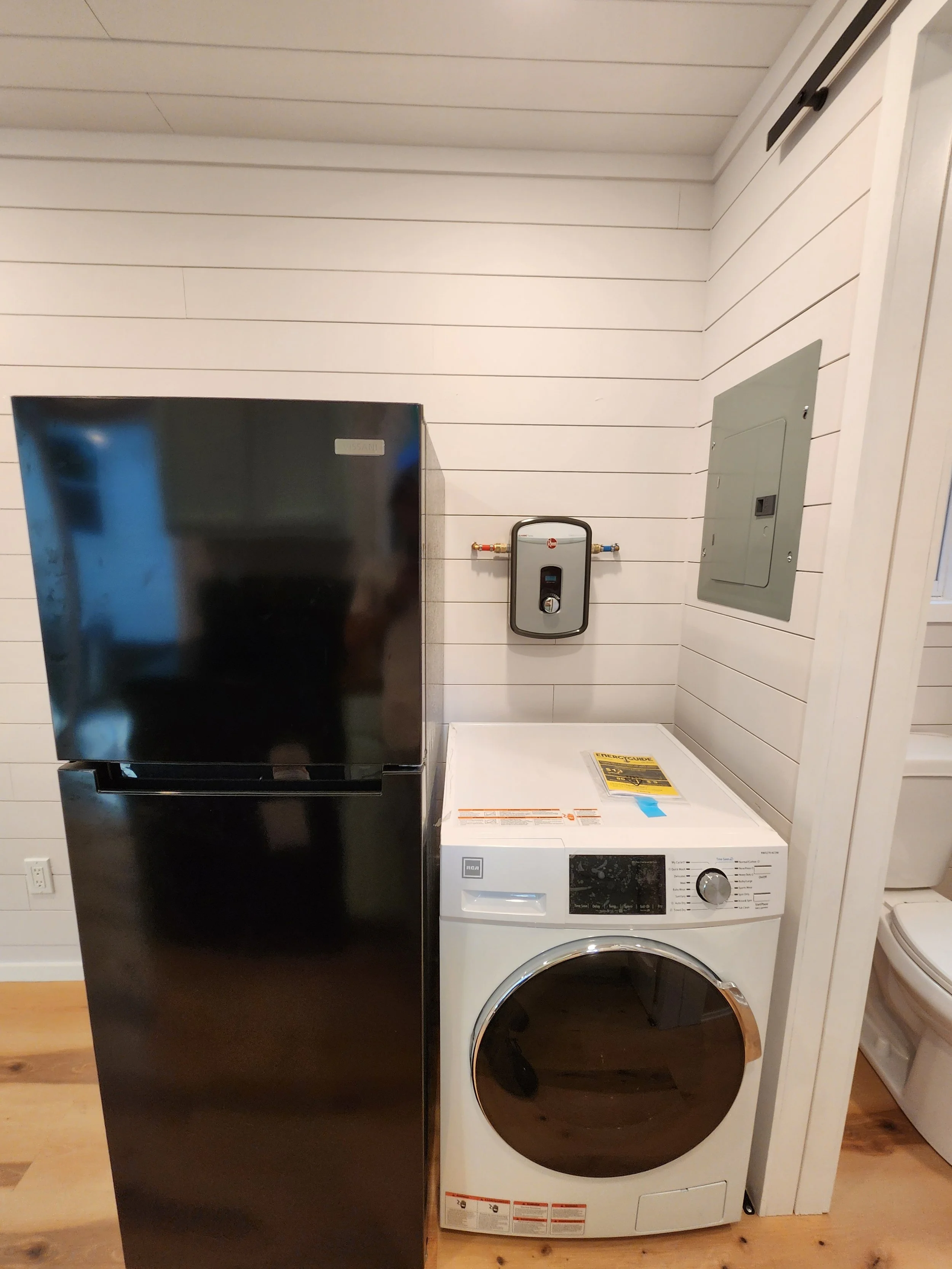 A laundry area with a black refrigerator on the left and a white front-load washing machine on the right. There is an electrical panel on the wall behind.