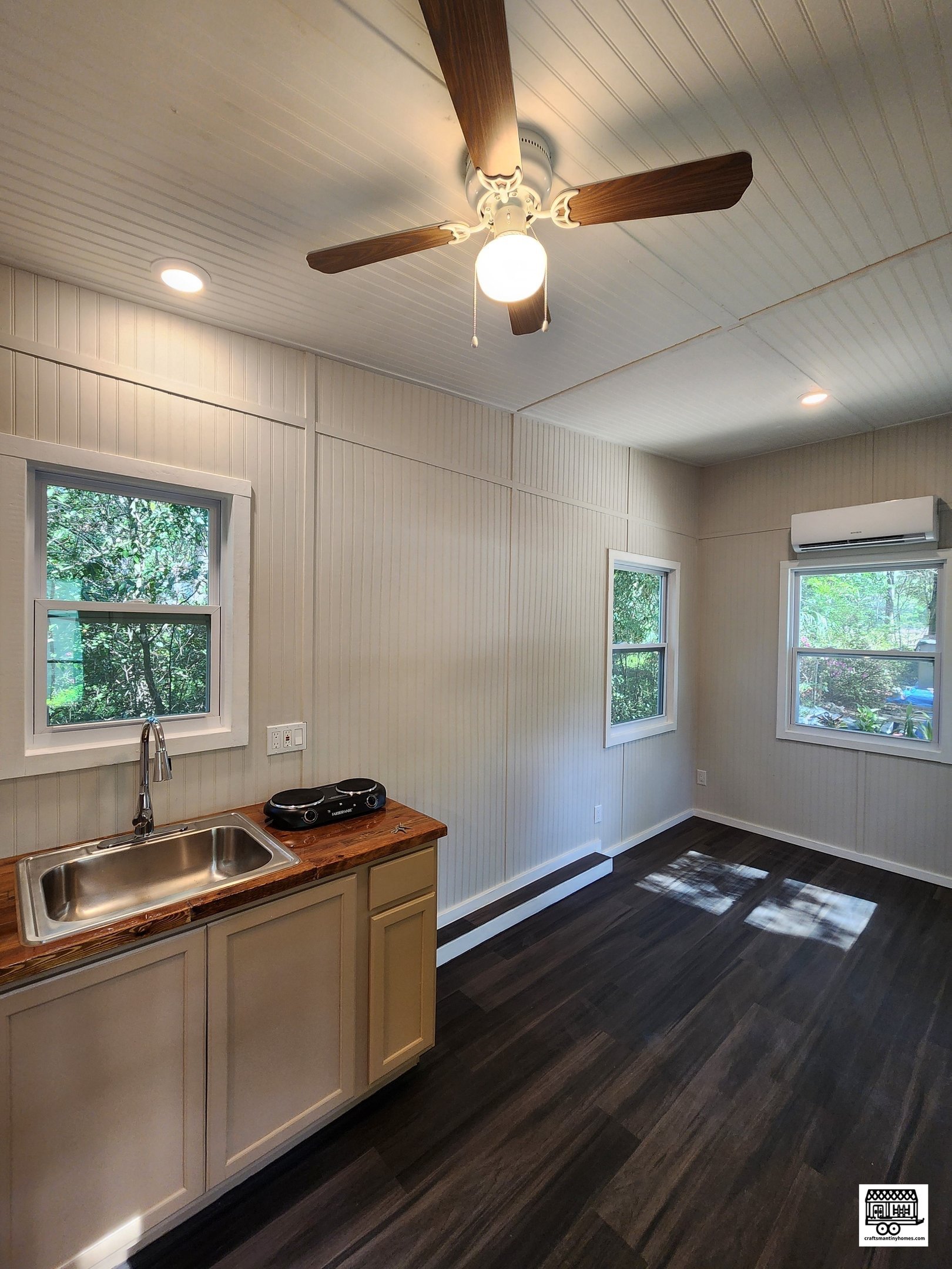 Interior of a small room with white paneled walls and ceiling, wooden flooring, two windows showing trees outside, a ceiling fan with light, a mounted air conditioning unit, a countertop with a sink, and a portable stovetop.