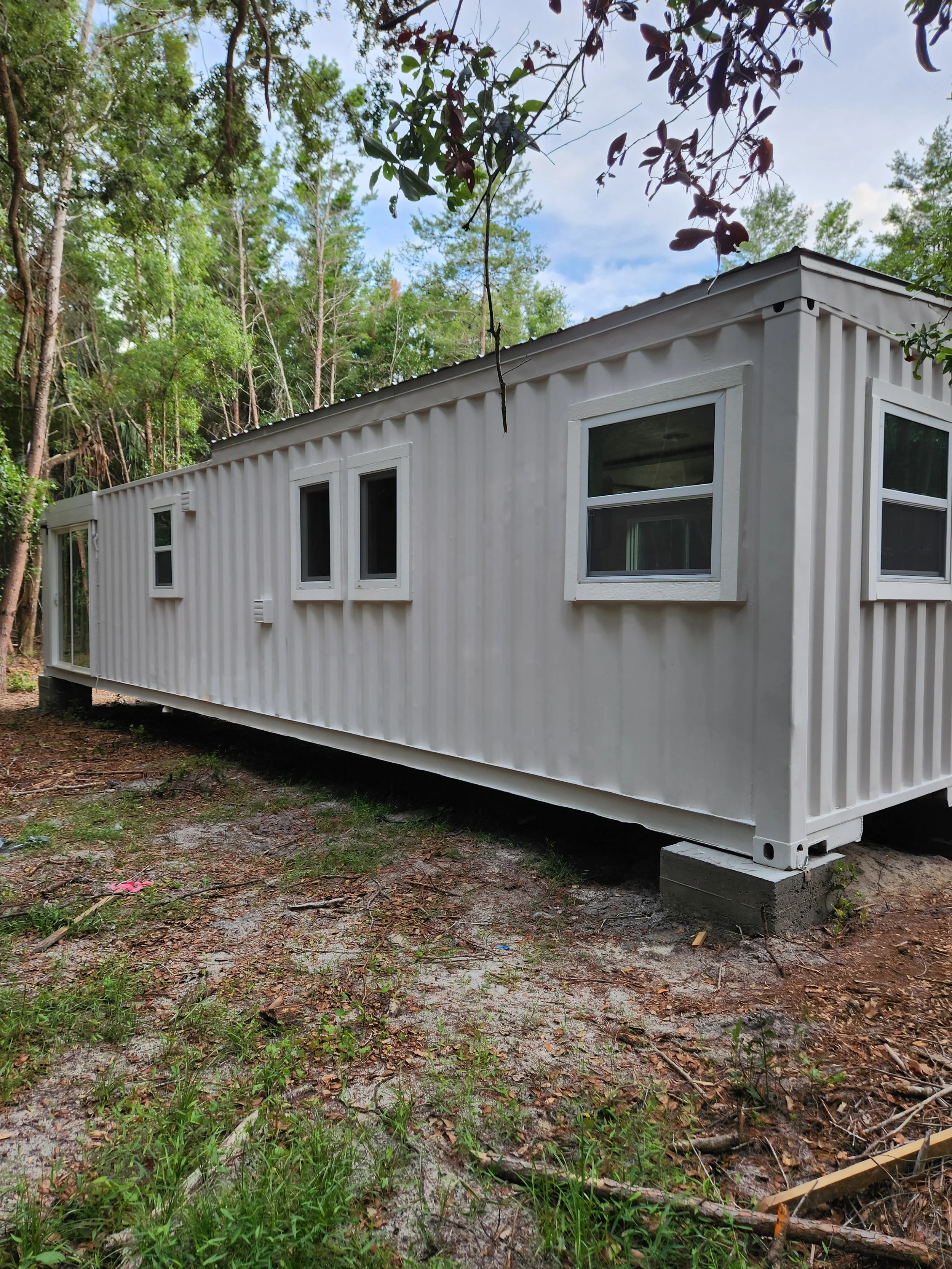 A white shipping container home with multiple windows, elevated on concrete blocks, situated in a wooded outdoor area with trees and a partly cloudy sky.