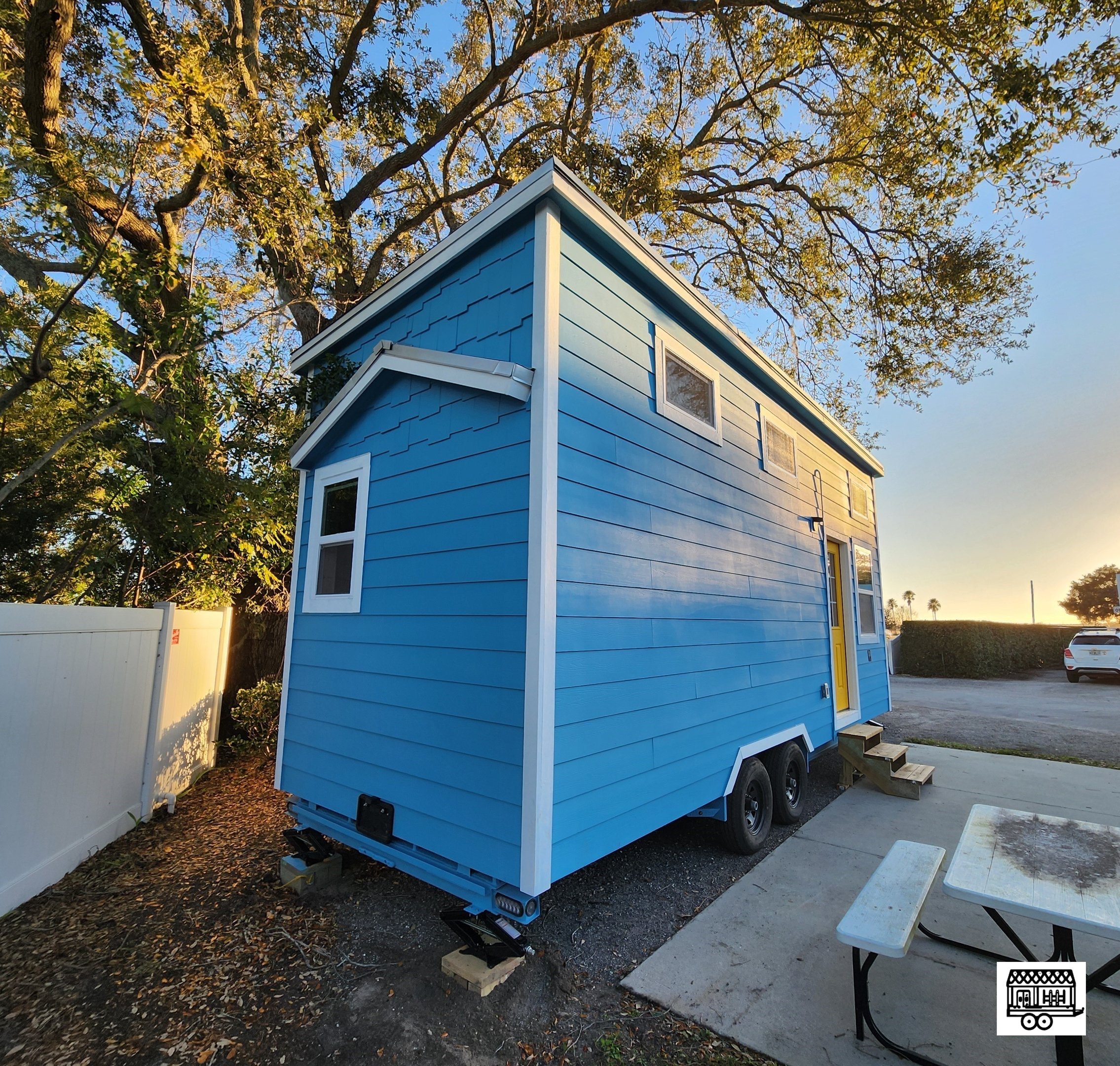 A small, blue, two-story tiny house on wheels with white trim and multiple small windows, located outdoors at sunset with trees and a parking lot nearby.
