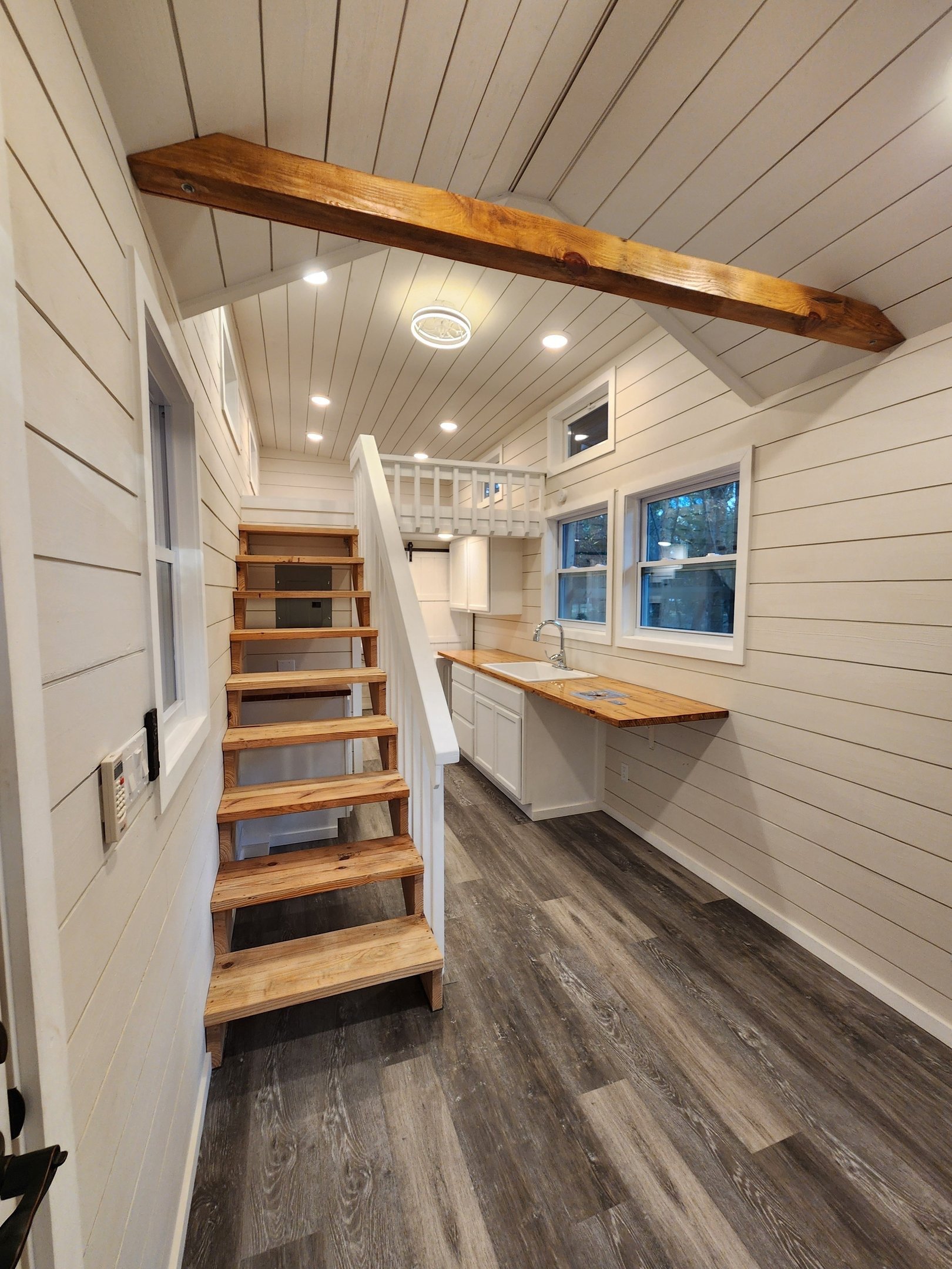 A small kitchen with white shiplap walls and ceiling, wooden flooring, a wooden working surface, and a staircase leading to a loft area. There are three windows, a sink, and a mix of recessed and ceiling light fixtures.