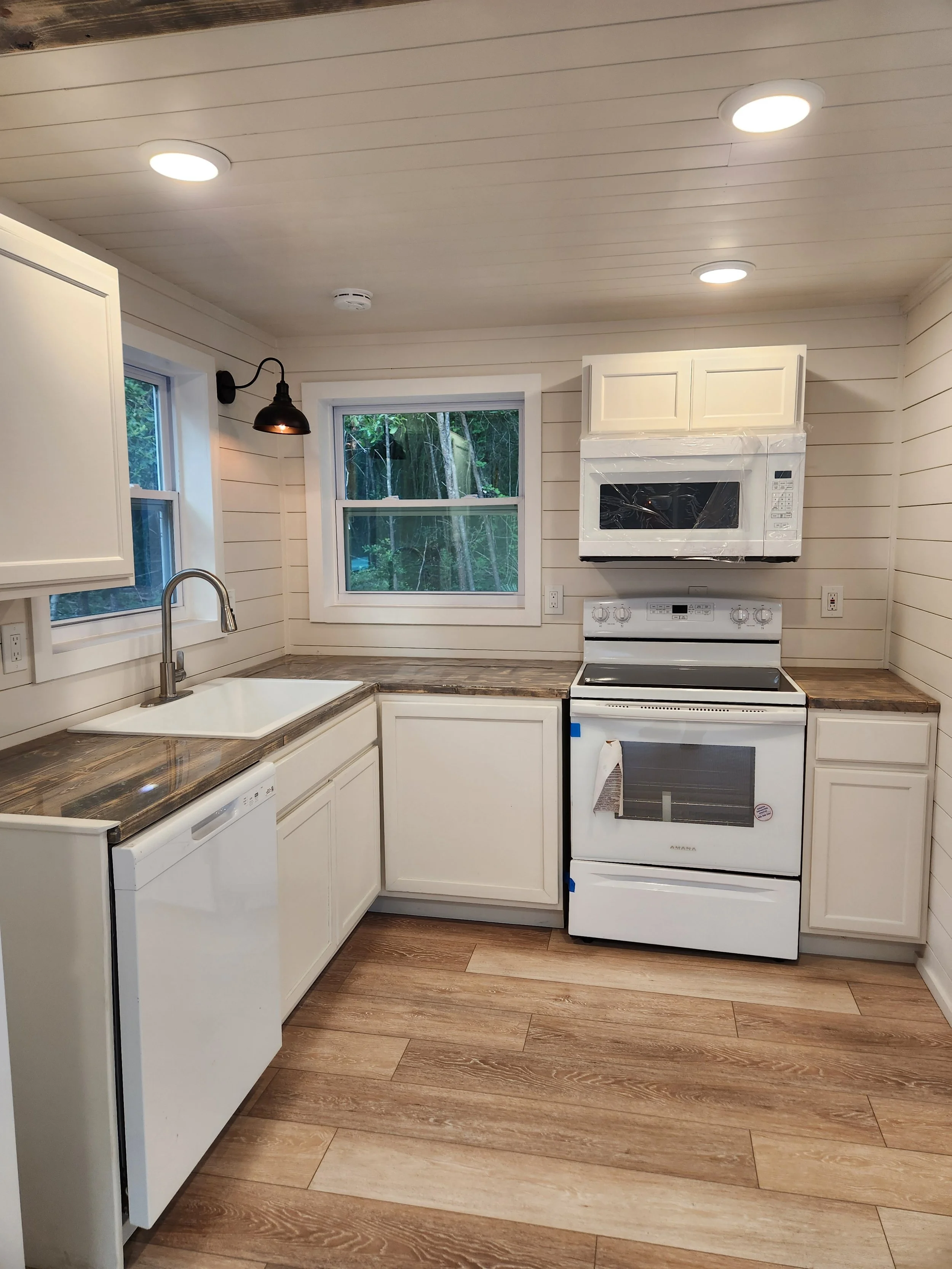 Kitchen with white cabinets, brown granite countertops, a black wall-mounted lamp, a window with a view of green trees, a white sink, a dishwasher, a stove, and a microwave.