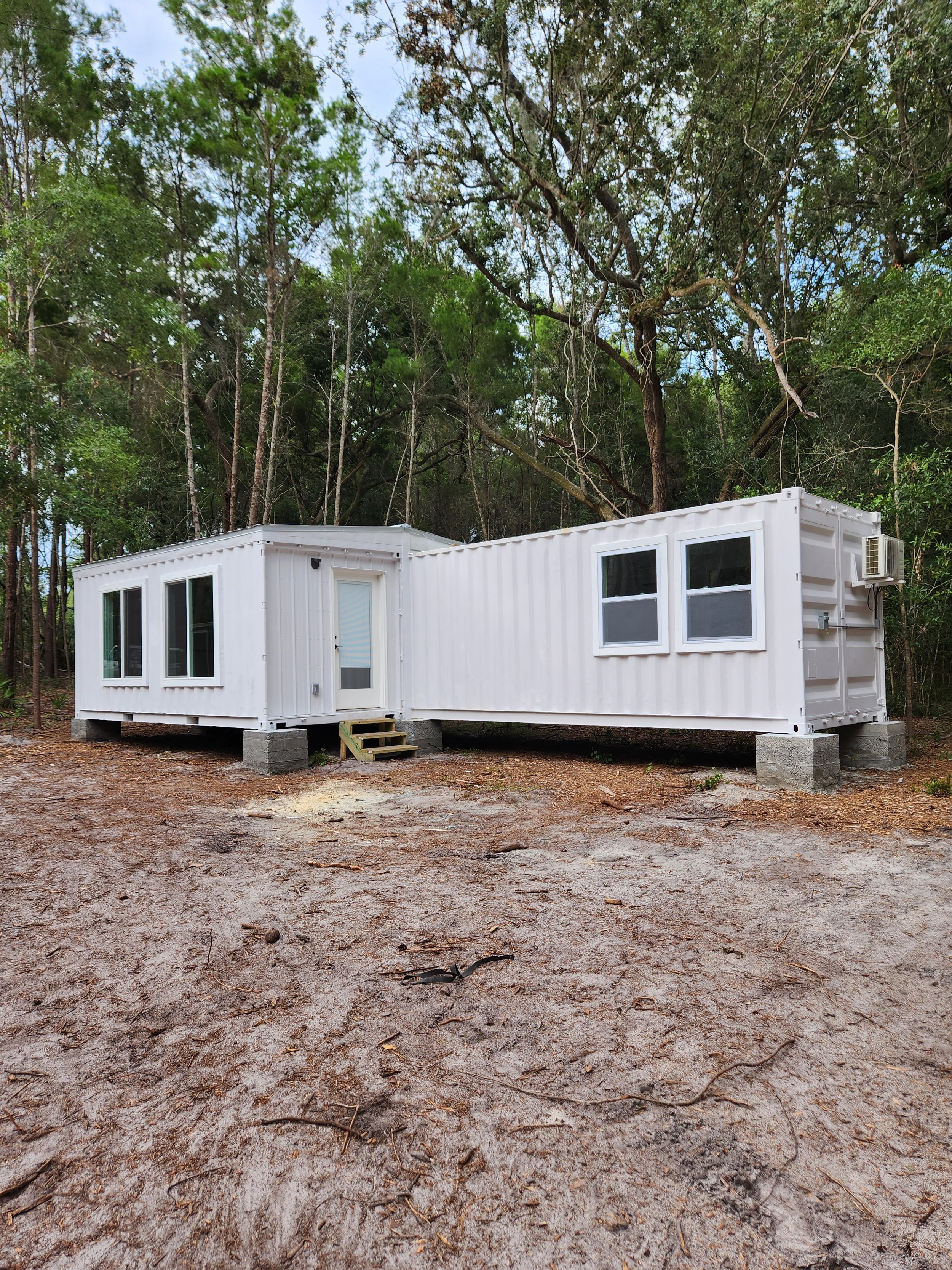 Two white shipping container homes elevated on concrete blocks in a wooded area with trees in the background.