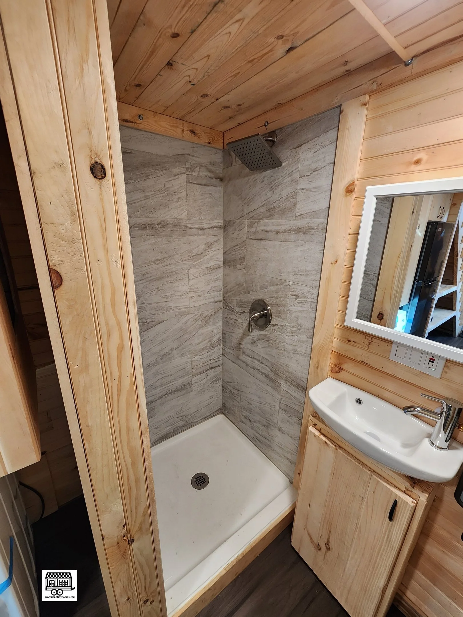 Small bathroom with wooden walls and ceiling, a shower with gray tile walls, a square showerhead, and a single faucet; a small white sink with a modern faucet, a mirror, and electrical outlets