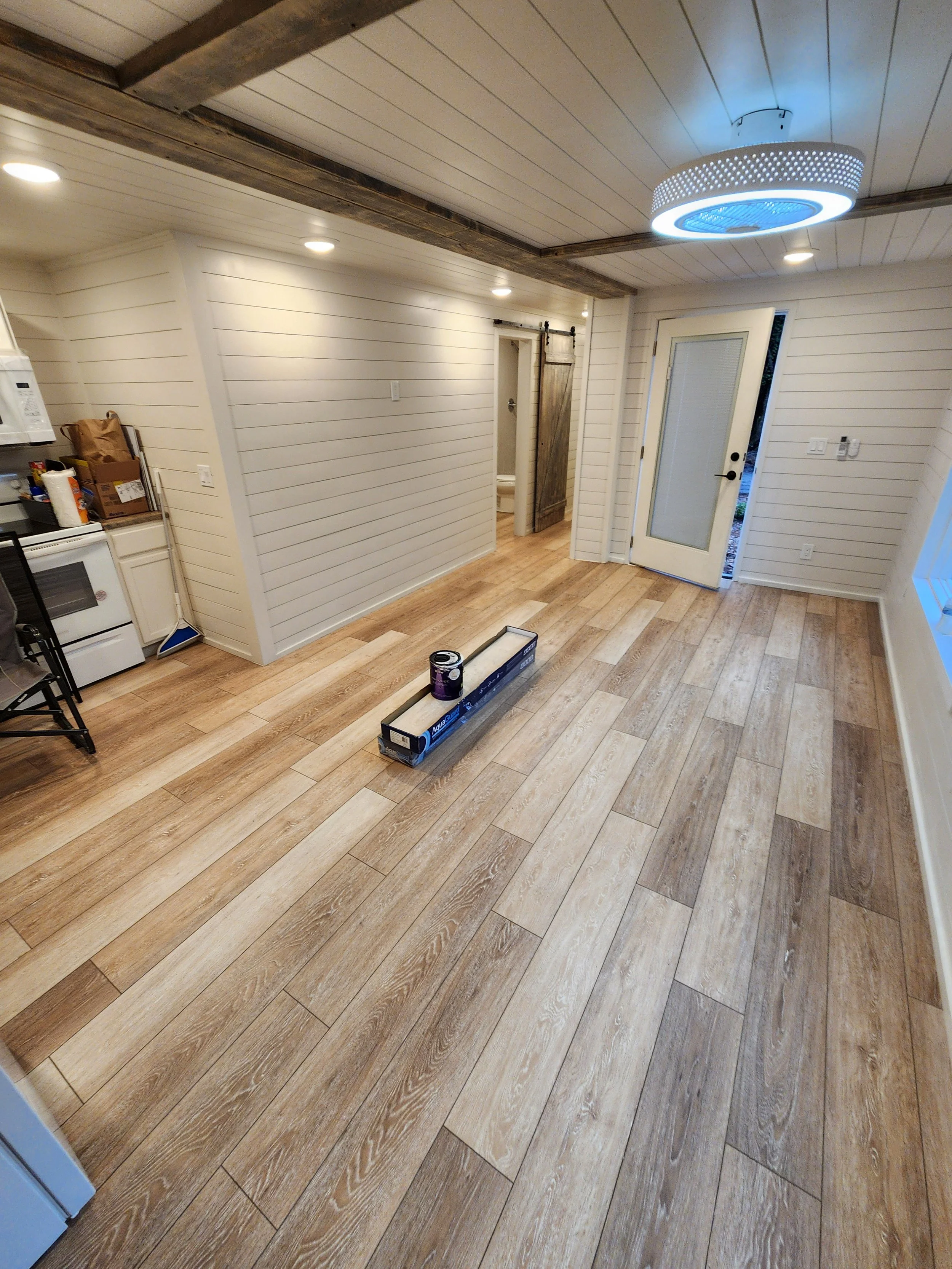 Room with wood flooring, white paneled walls, modern ceiling light, small kitchen area, and a door leading outside. A box of flooring and a can of paint are on the floor.