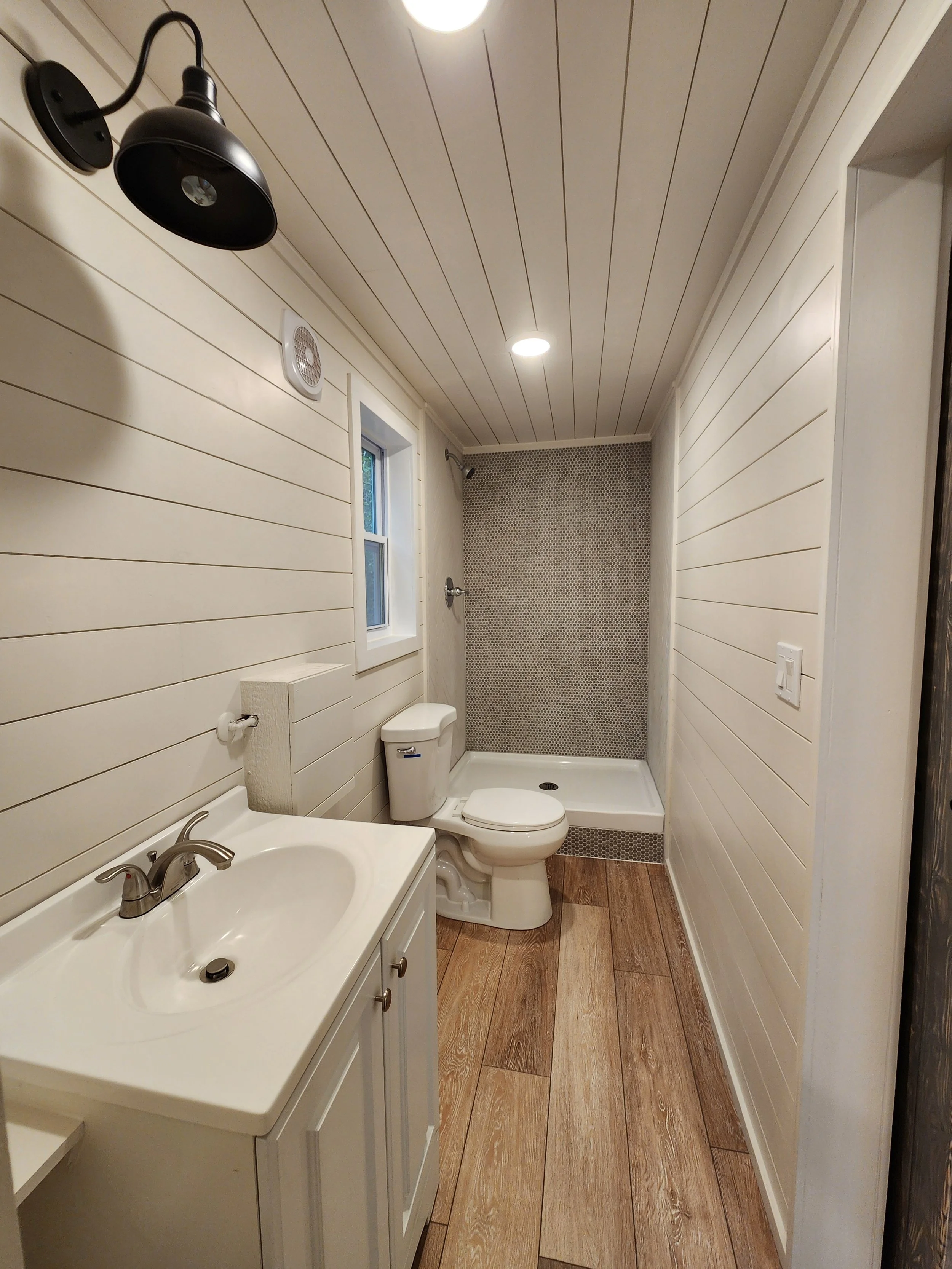 A small bathroom with wood plank flooring, white shiplap walls and ceiling, a small window, black wall sconce, toilet, white vanity with sink, and a walk-in shower with small mosaic tiles on the back wall.