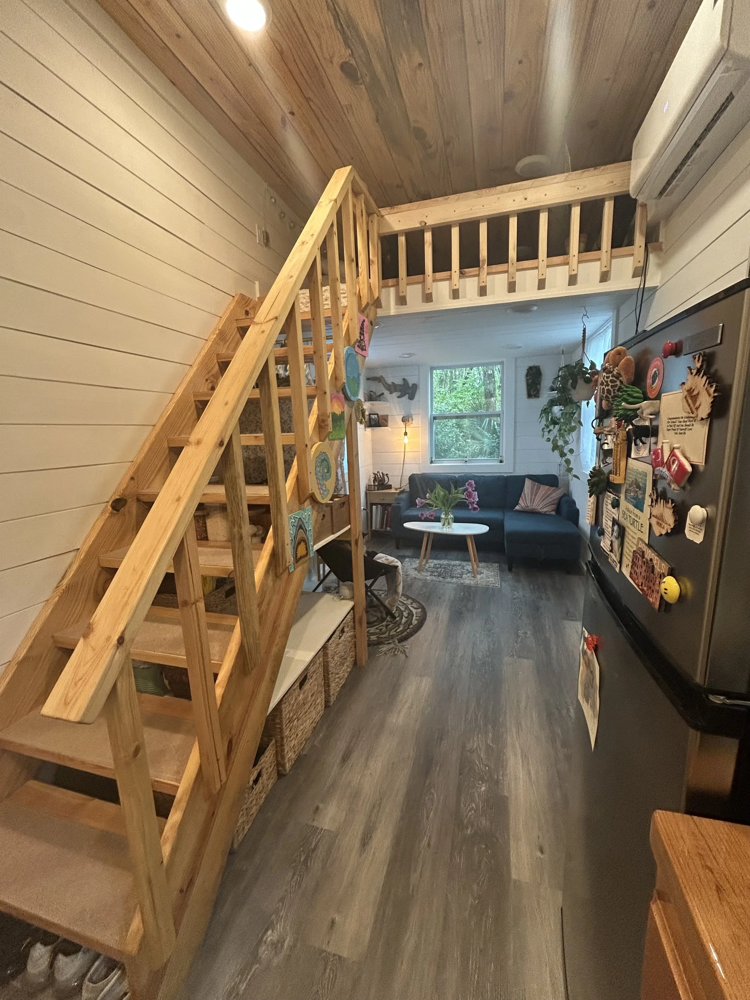 Cozy tiny home living room with loft stairs, a blue sectional sofa, and natural light — modern small-space design by Crafted Tiny Homes USA for minimalist and sustainable living.