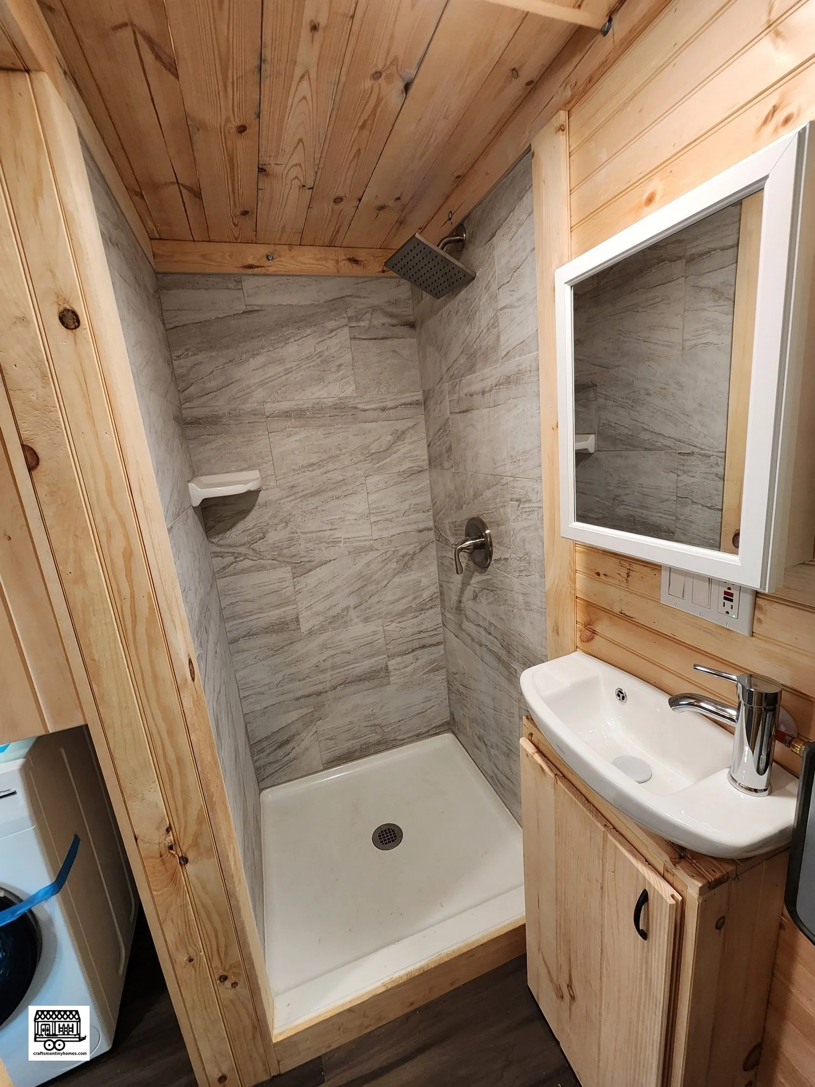 Small bathroom with a shower area featuring gray marble tiles, a wooden ceiling and walls, a small white sink with a chrome faucet, a mirror, and a washing machine in the corner.