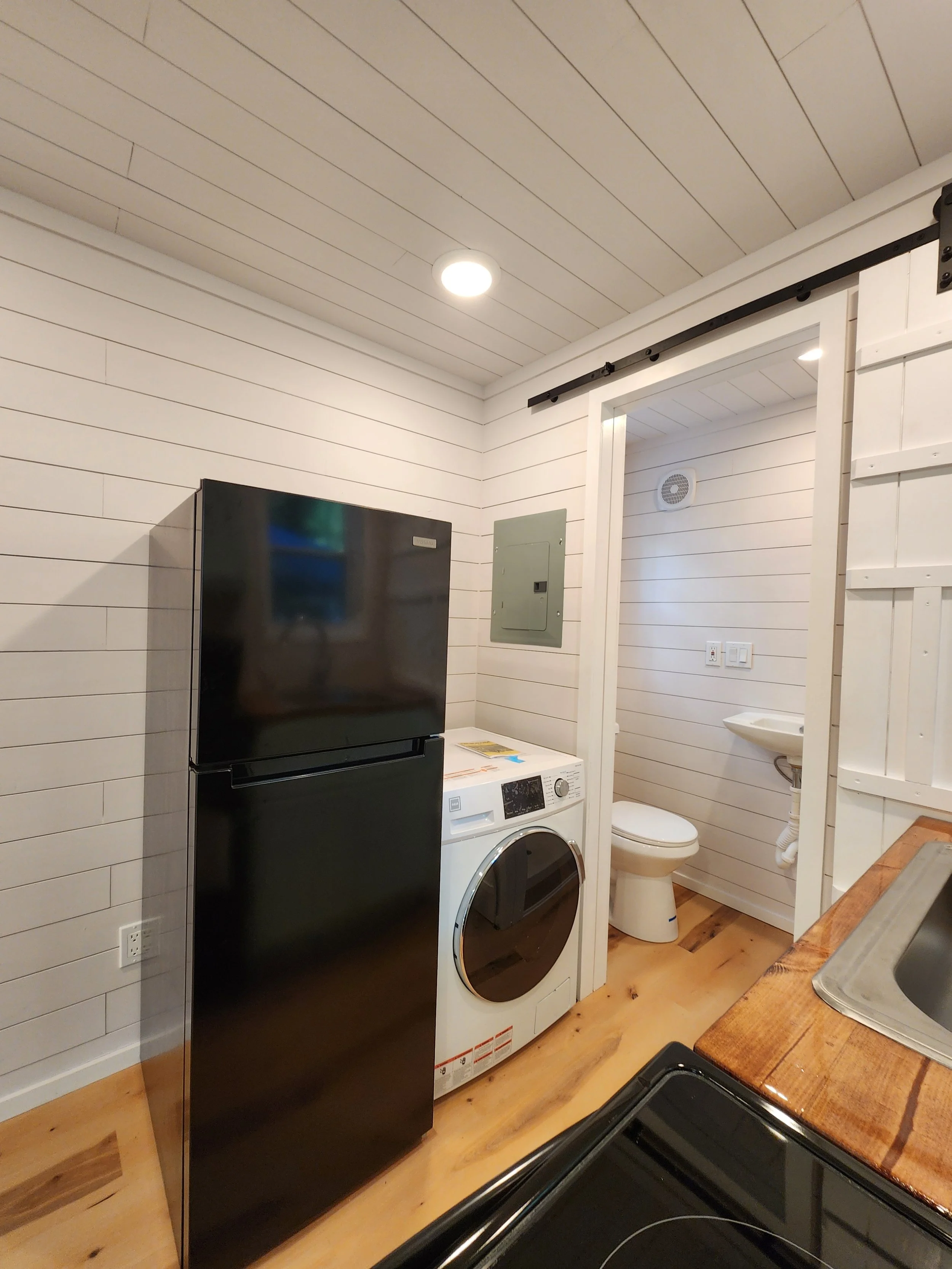 A small laundry room with a black refrigerator, a washing machine, and a small bathroom with a toilet and a sink.