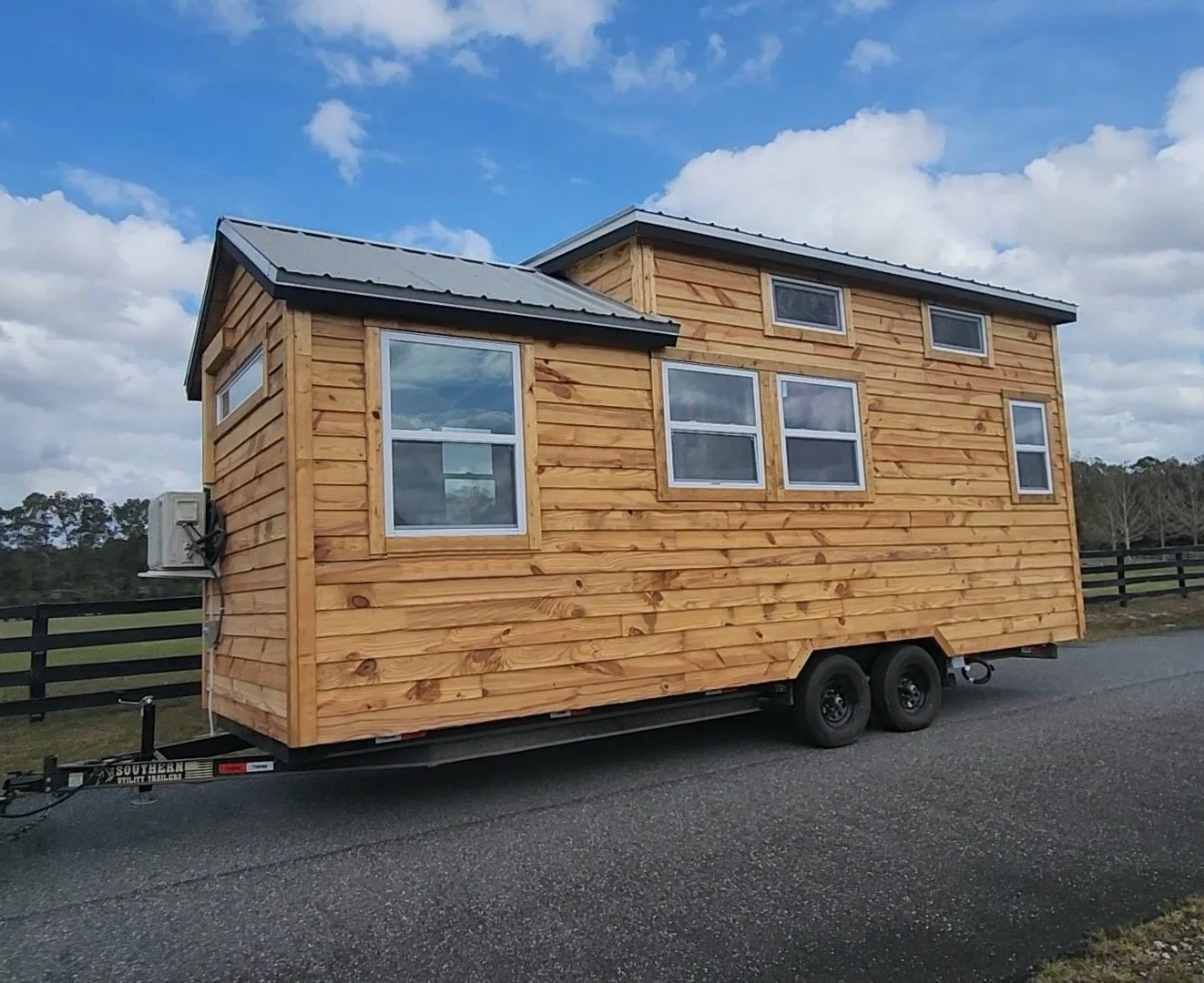 A tiny house on wheels with wood siding and multiple windows parked on a paved road under a cloudy sky.