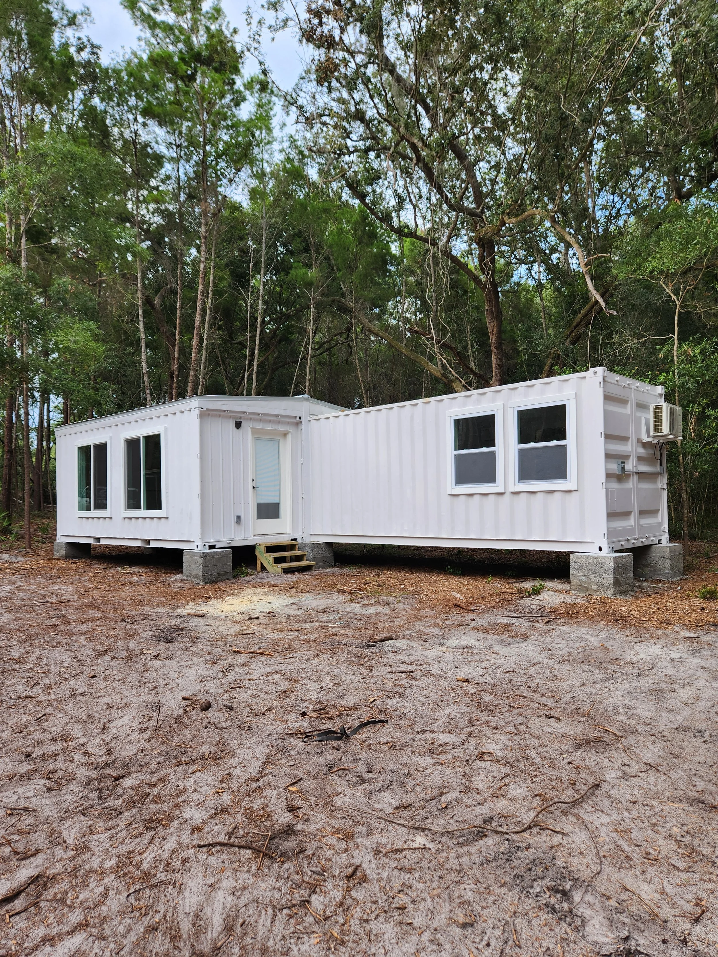 A white tiny house built with shipping containers, elevated on concrete blocks, with trees and forest in the background.