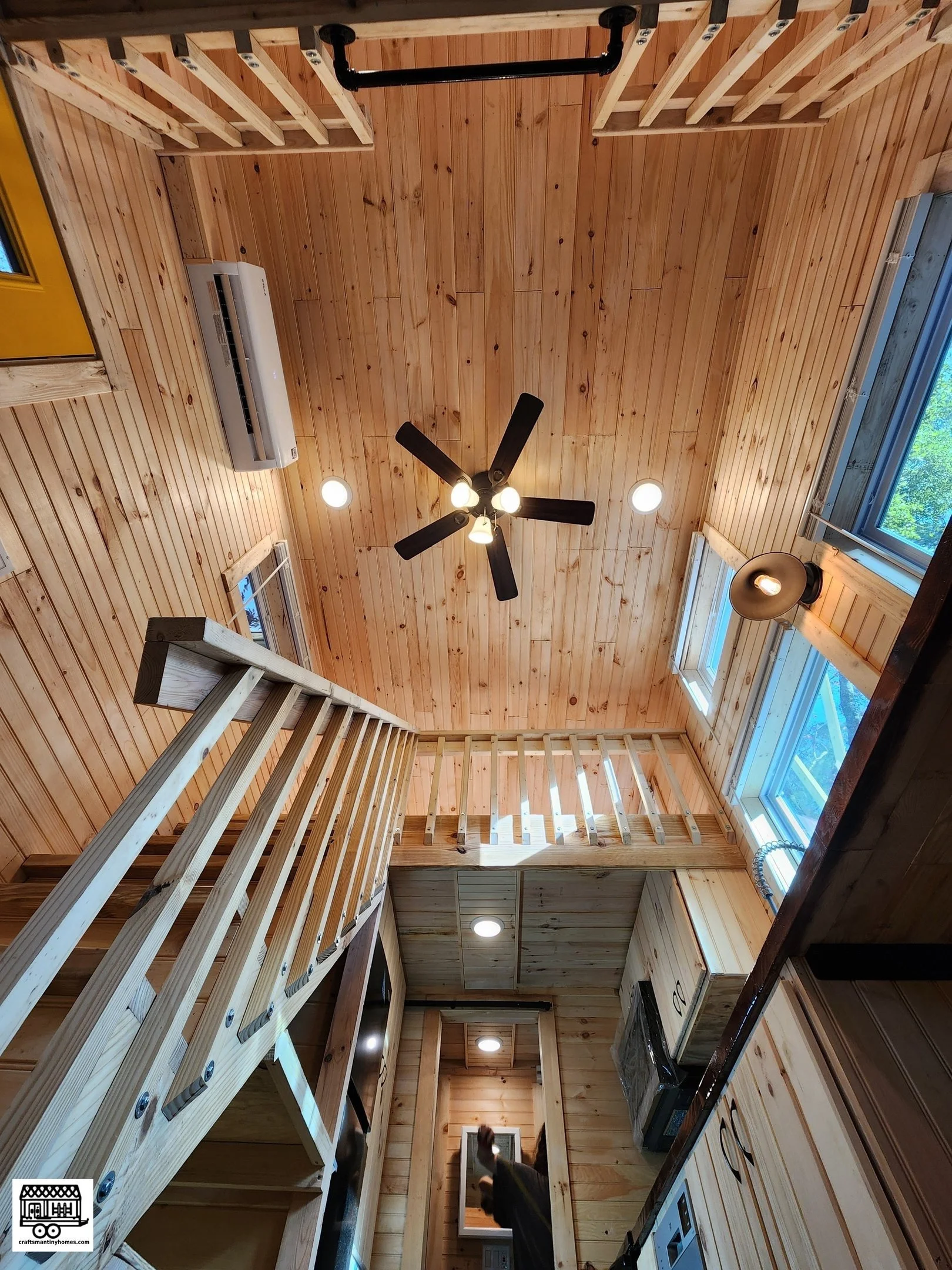 Interior view of a wooden cabin with vaulted ceiling, ceiling fans, skylights, and an open loft with a railing, featuring kitchen appliances and cabinets.