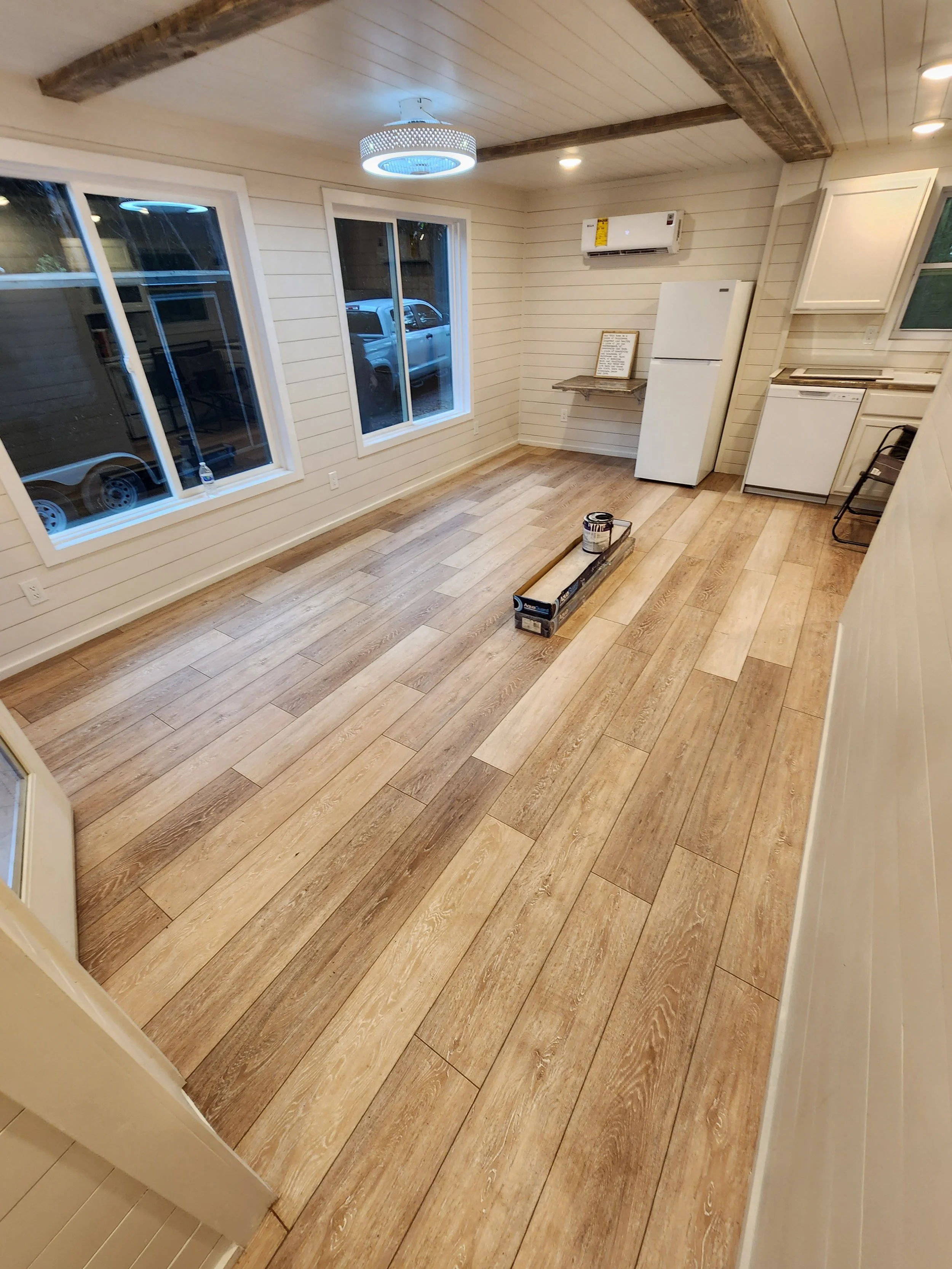 Room with new wood-look flooring, three large windows, white walls, a ceiling light, and kitchen appliances including a refrigerator and dishwasher, with some tools and a paint can on the floor.