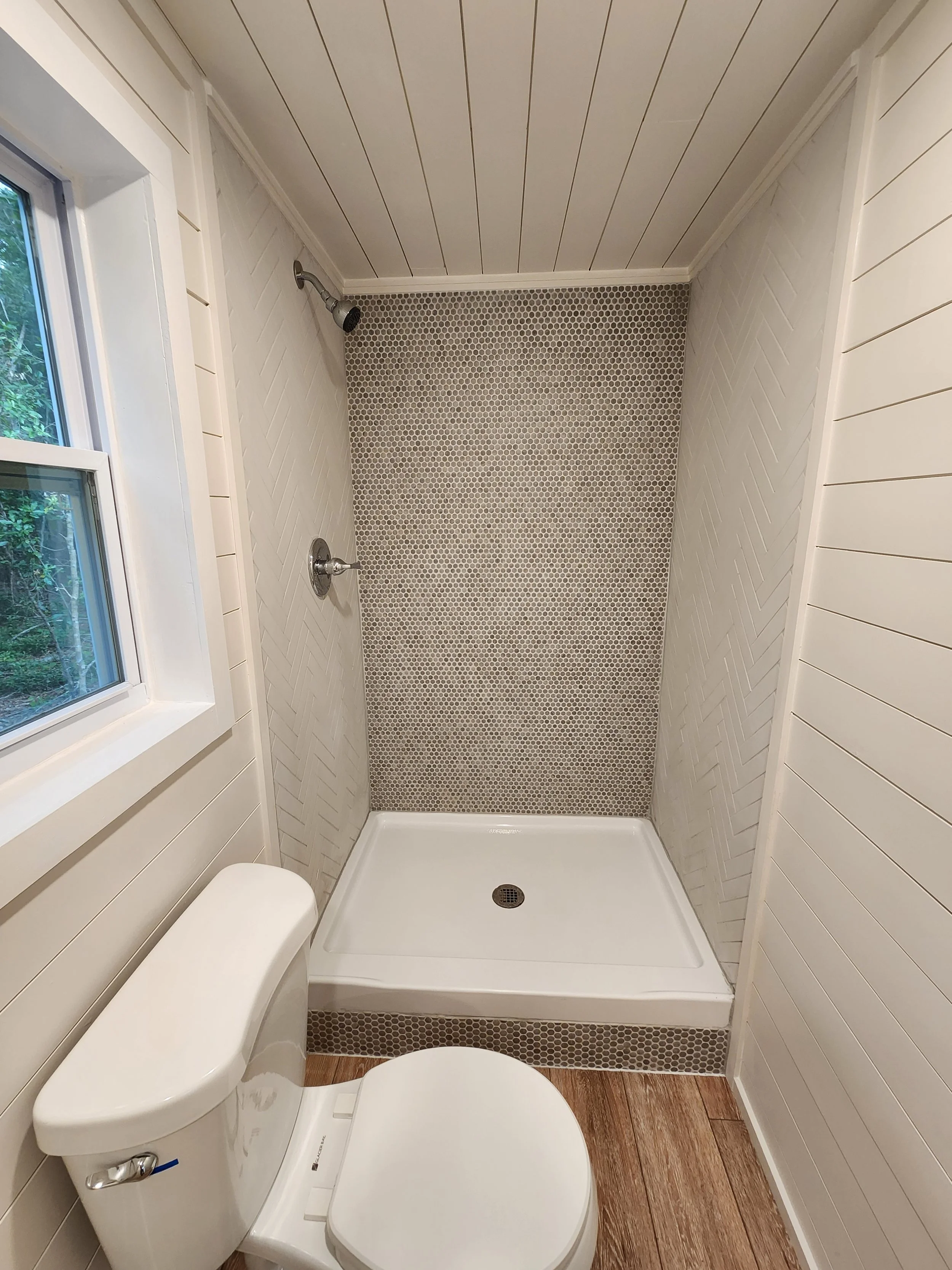 Small bathroom with a toilet next to a shower area with a built-in shower pan, white shiplap walls, and a window with greenery outside.