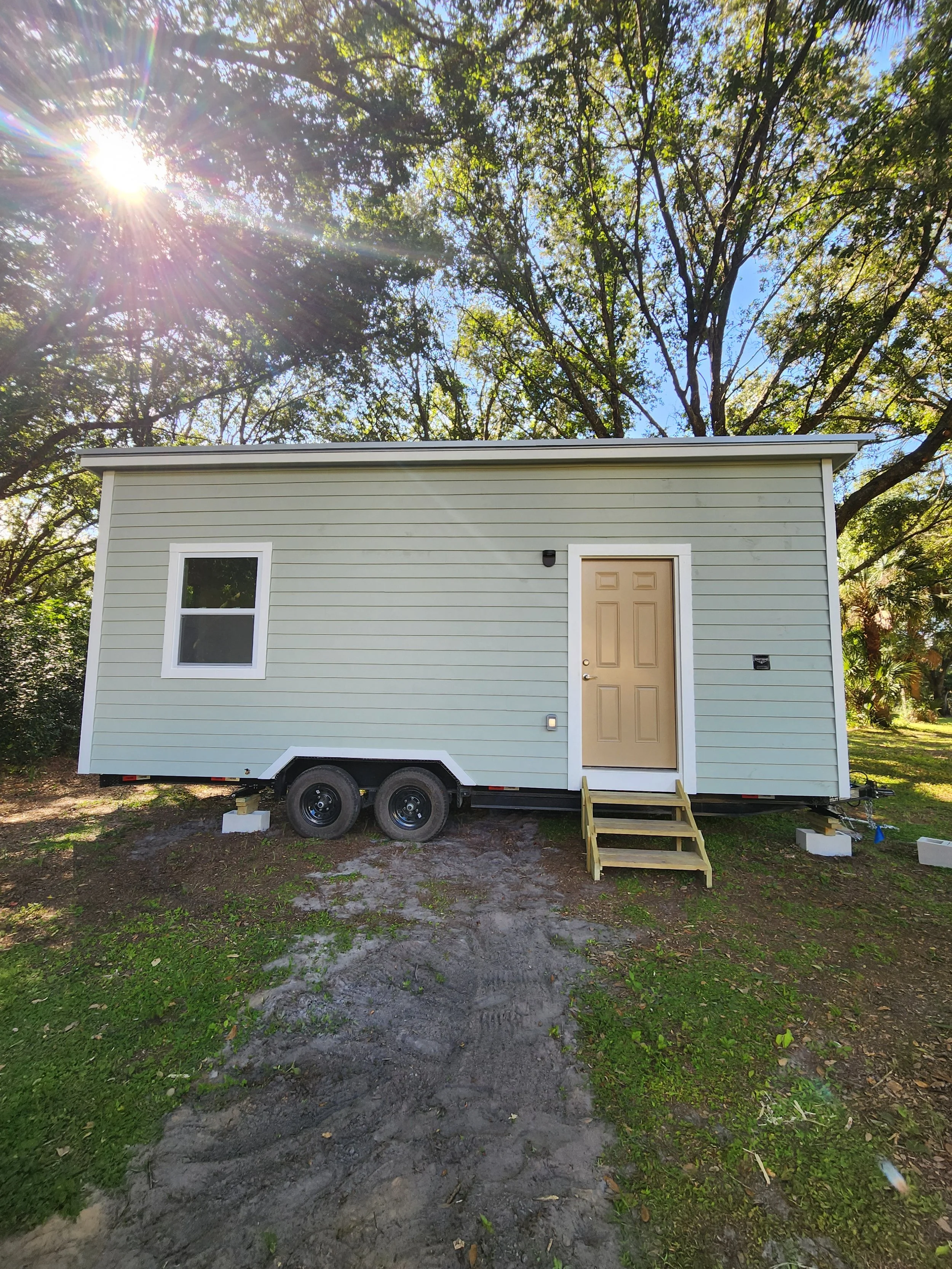 Tiny house on wheels with a beige door, a small window, and a set of stairs leading up to it, situated outdoors with trees and sunlight in the background.