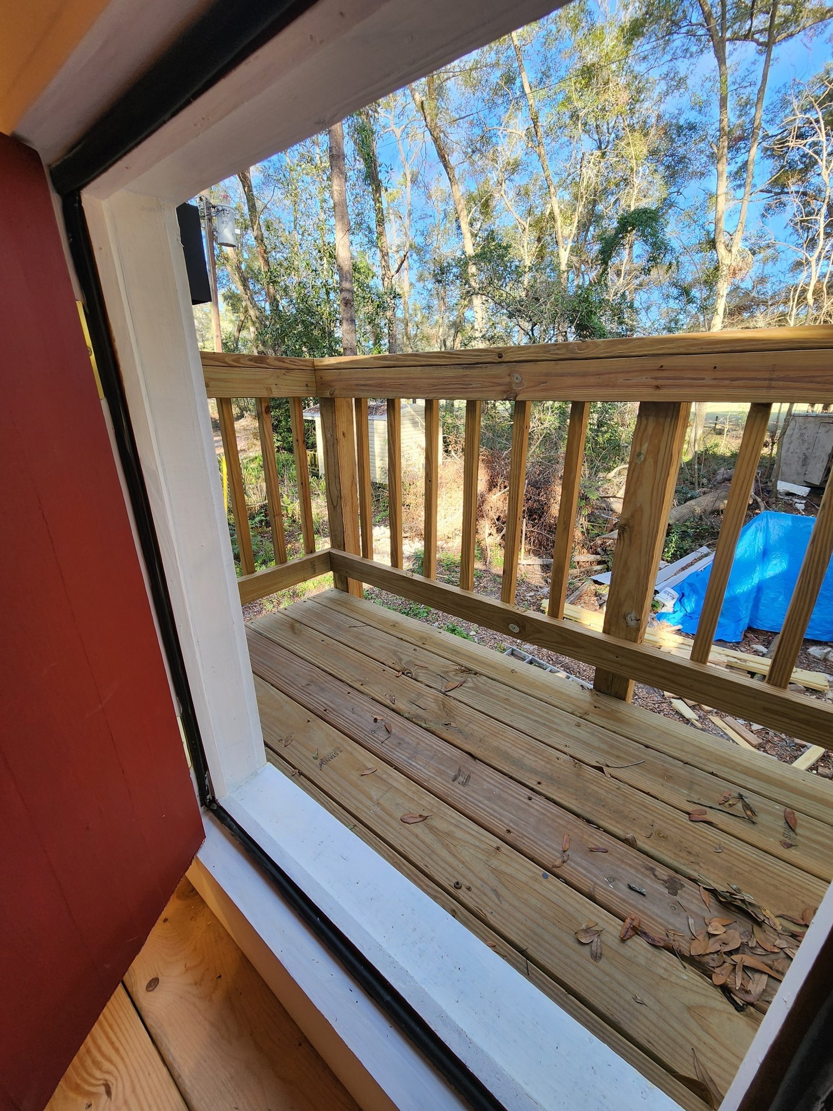 View from inside a building through a sliding glass door shows a small wooden balcony with a railing, overlooking a backyard with trees, a blue tarp, and construction materials.