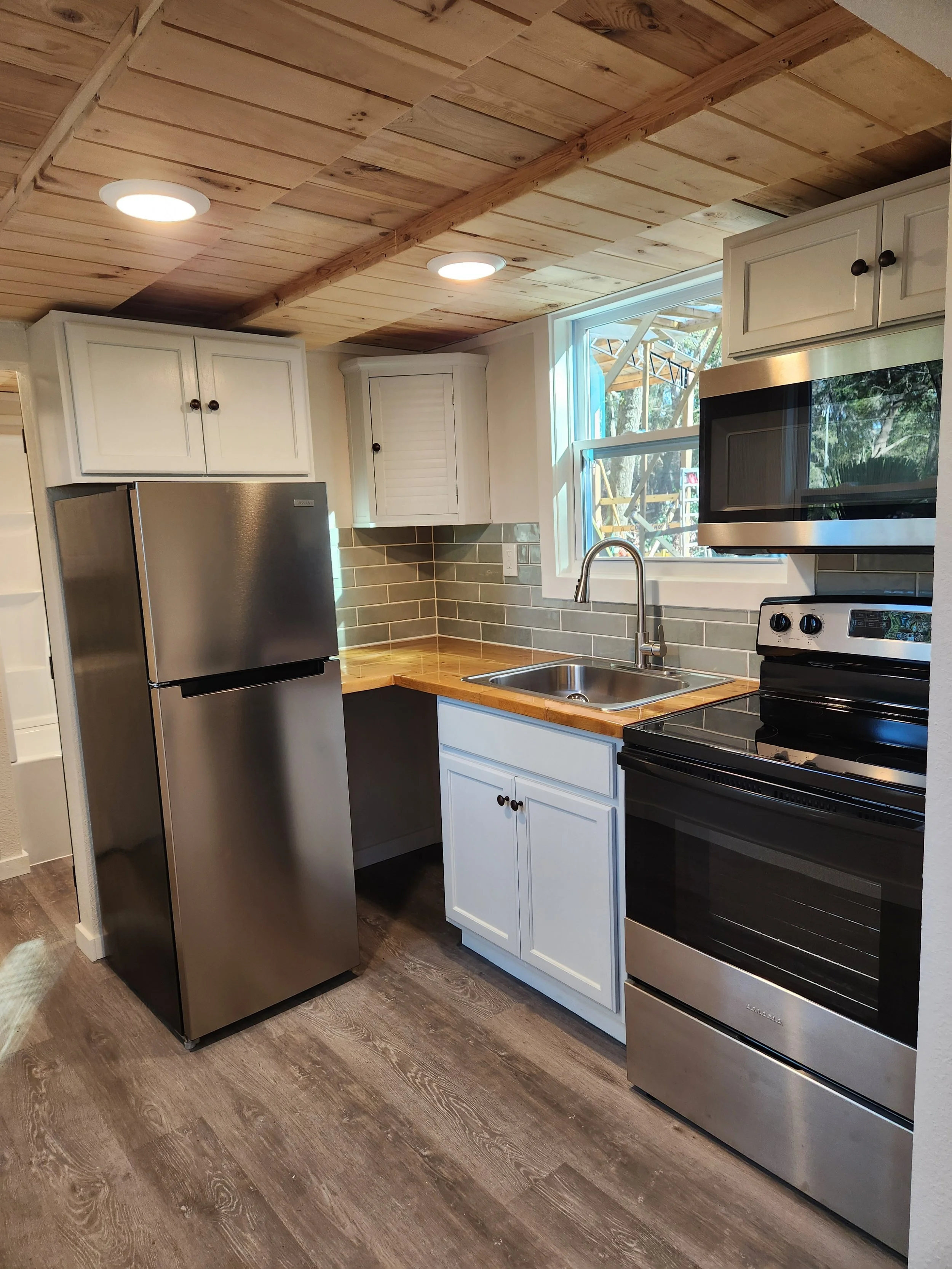 Kitchen with stainless steel refrigerator, oven, and microwave. White cabinetry, gray subway tile backsplash, wooden countertop around sink, and wooden plank ceiling. A window above the sink shows outdoor scenery.