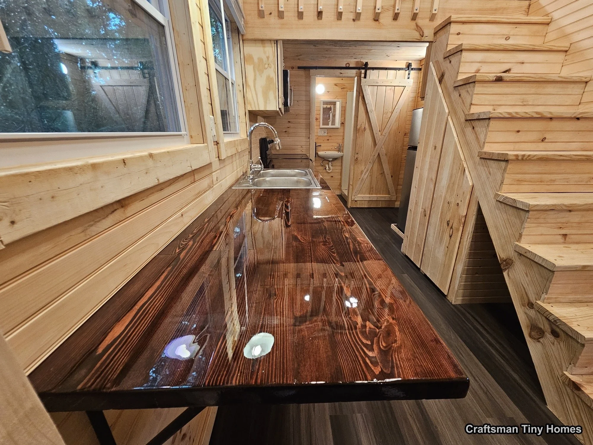 Interior of a tiny home with wood-paneled walls and staircase, kitchen area with a polished wooden countertop, sink, window with a view of trees, and a small bathroom in the background.