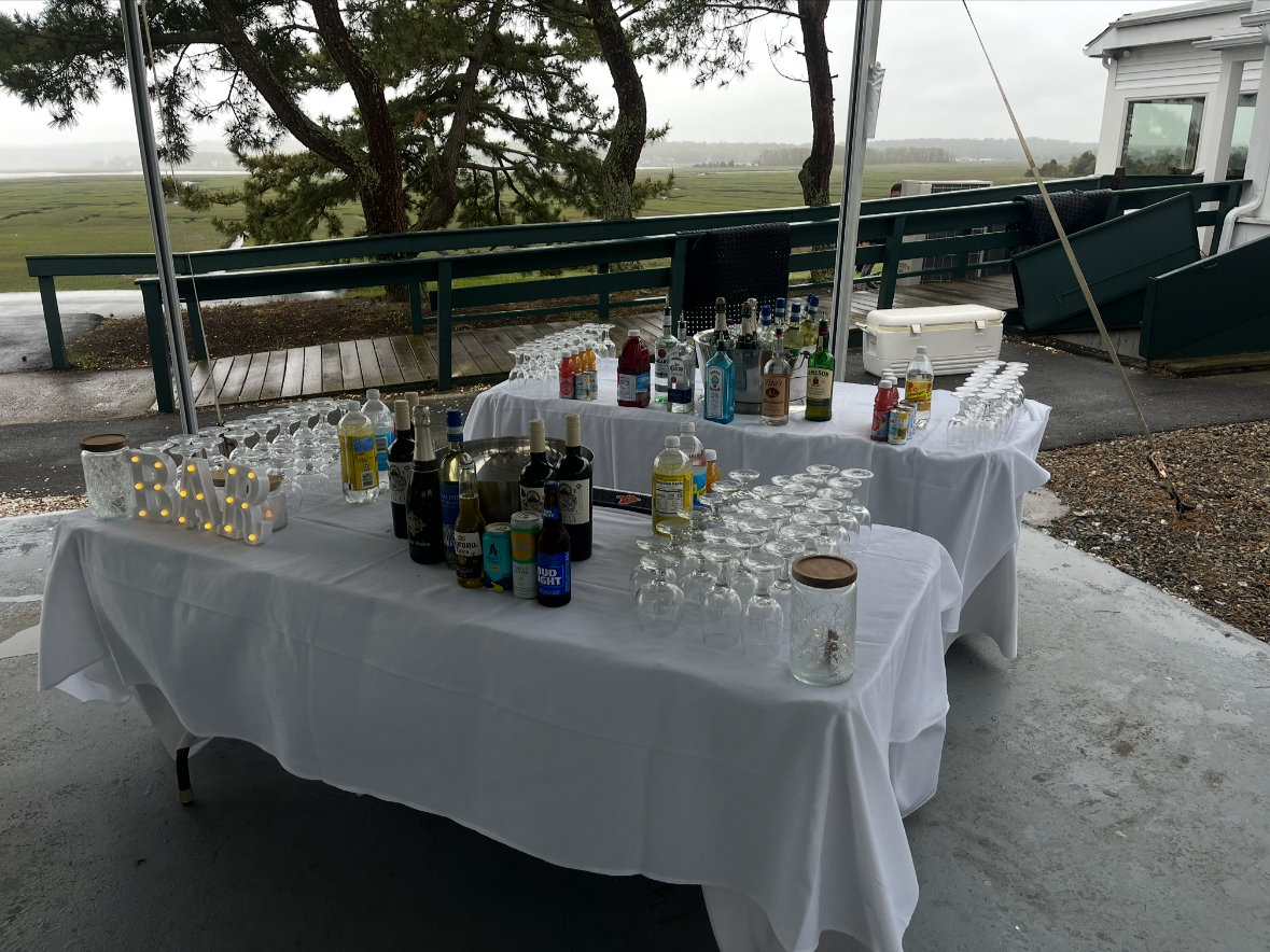 Two tables with white tablecloths set up outdoors with various bottles of alcohol, mixers, and glasses, likely for a bartending station at a gathering or party, with a view of a tree and open landscape in the background.
